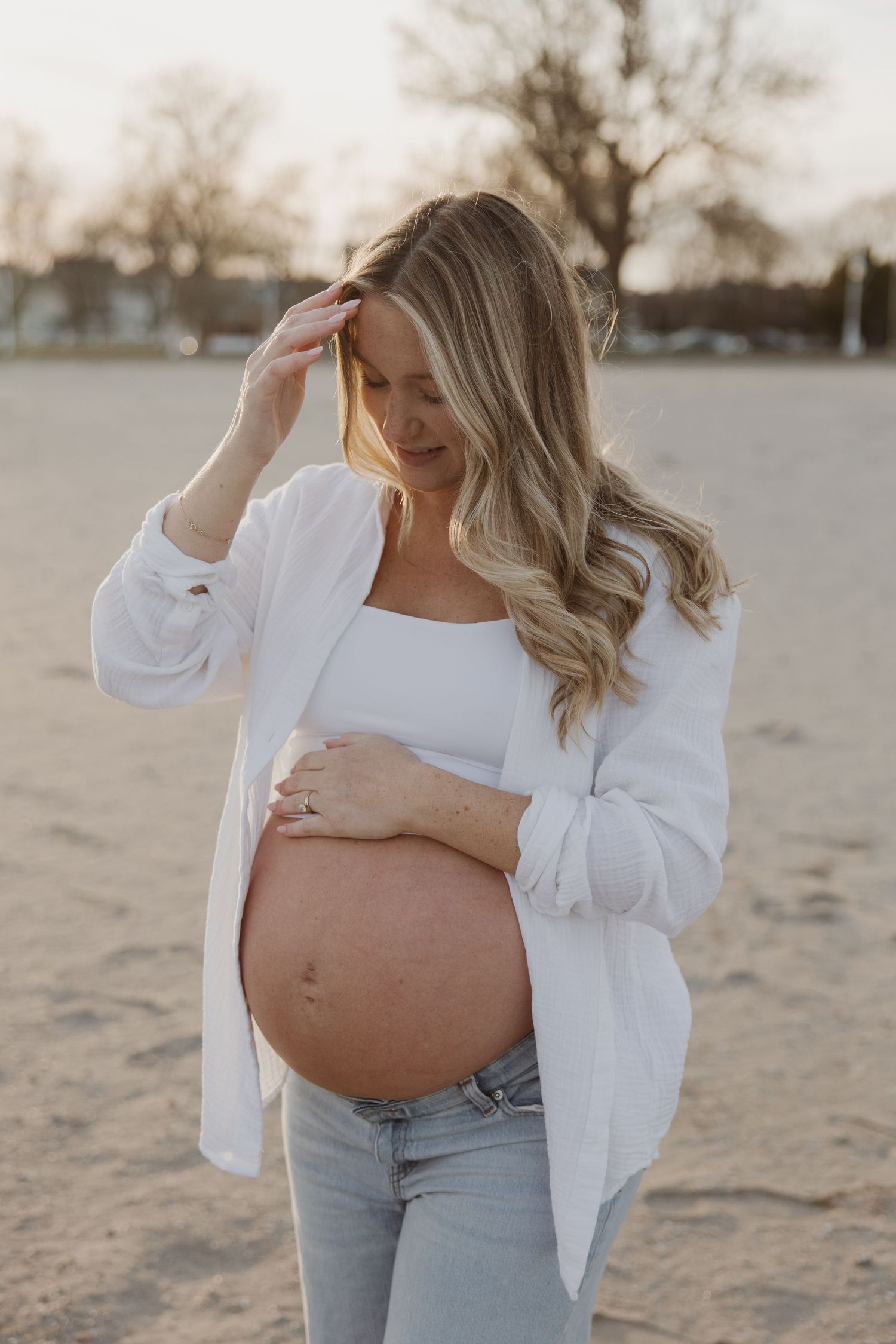 Pregnant woman in white shirt and jeans cradling her baby bump on a sandy beach at sunset.