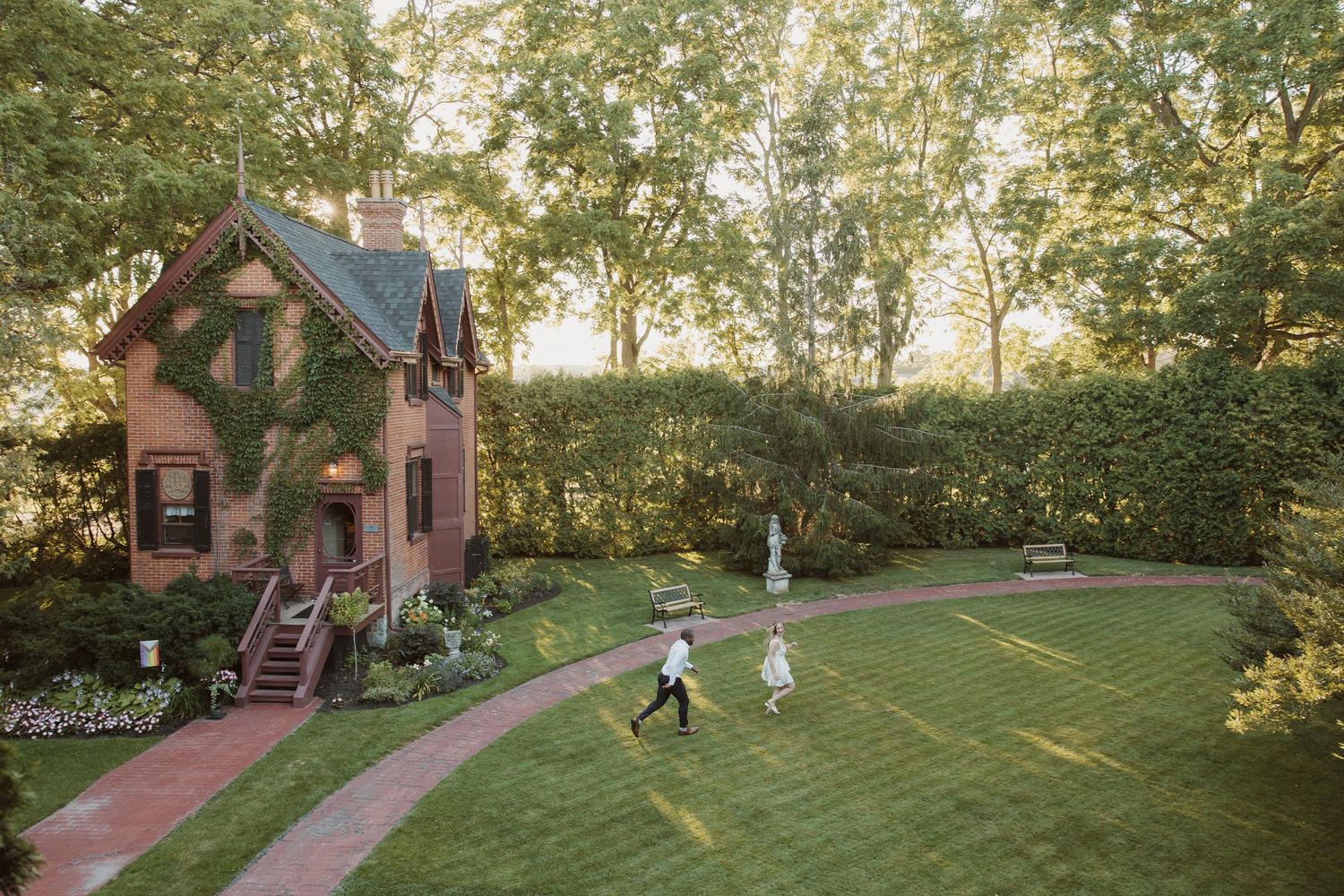 Aerial view of bride and groom walking across a lush garden lawn beside an ivy-covered brick cottage.