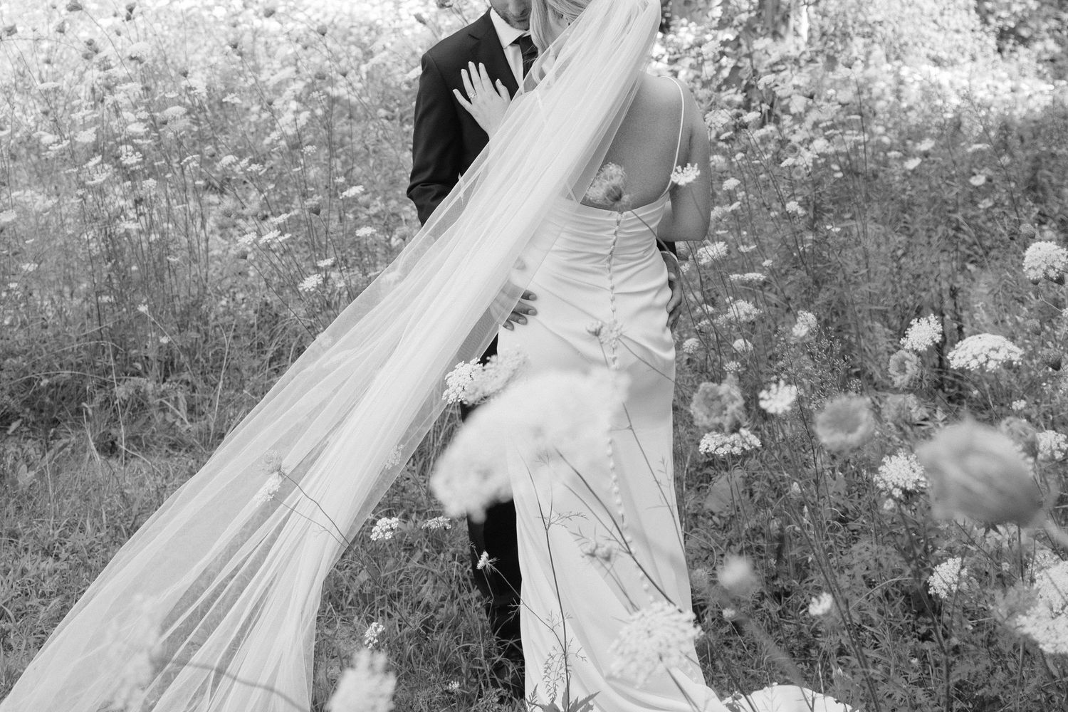 Black and white wedding photo of bride and groom in a wildflower field, veil flowing in the wind.