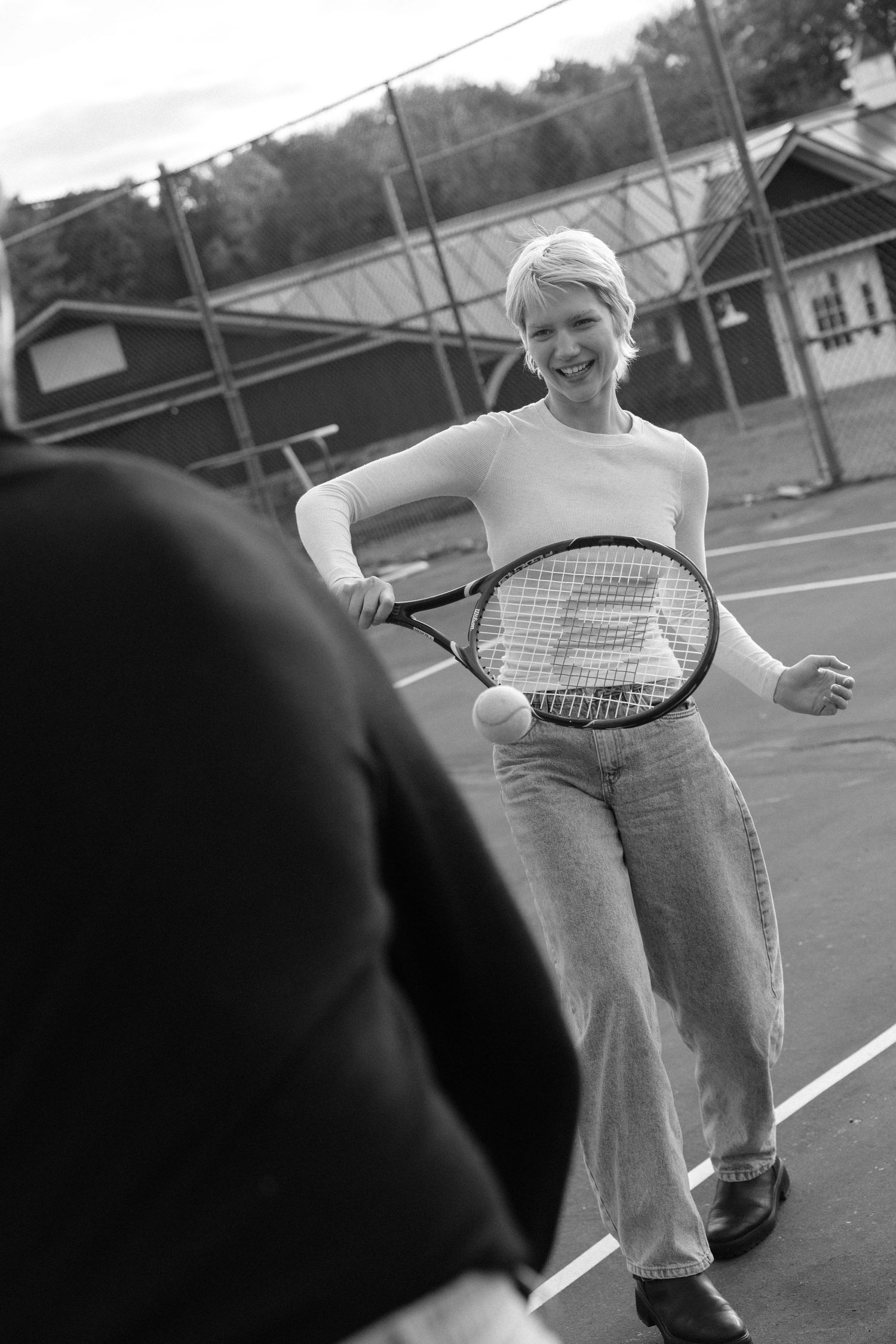 Woman smiling on tennis court holding racket with ball, black and white photography.