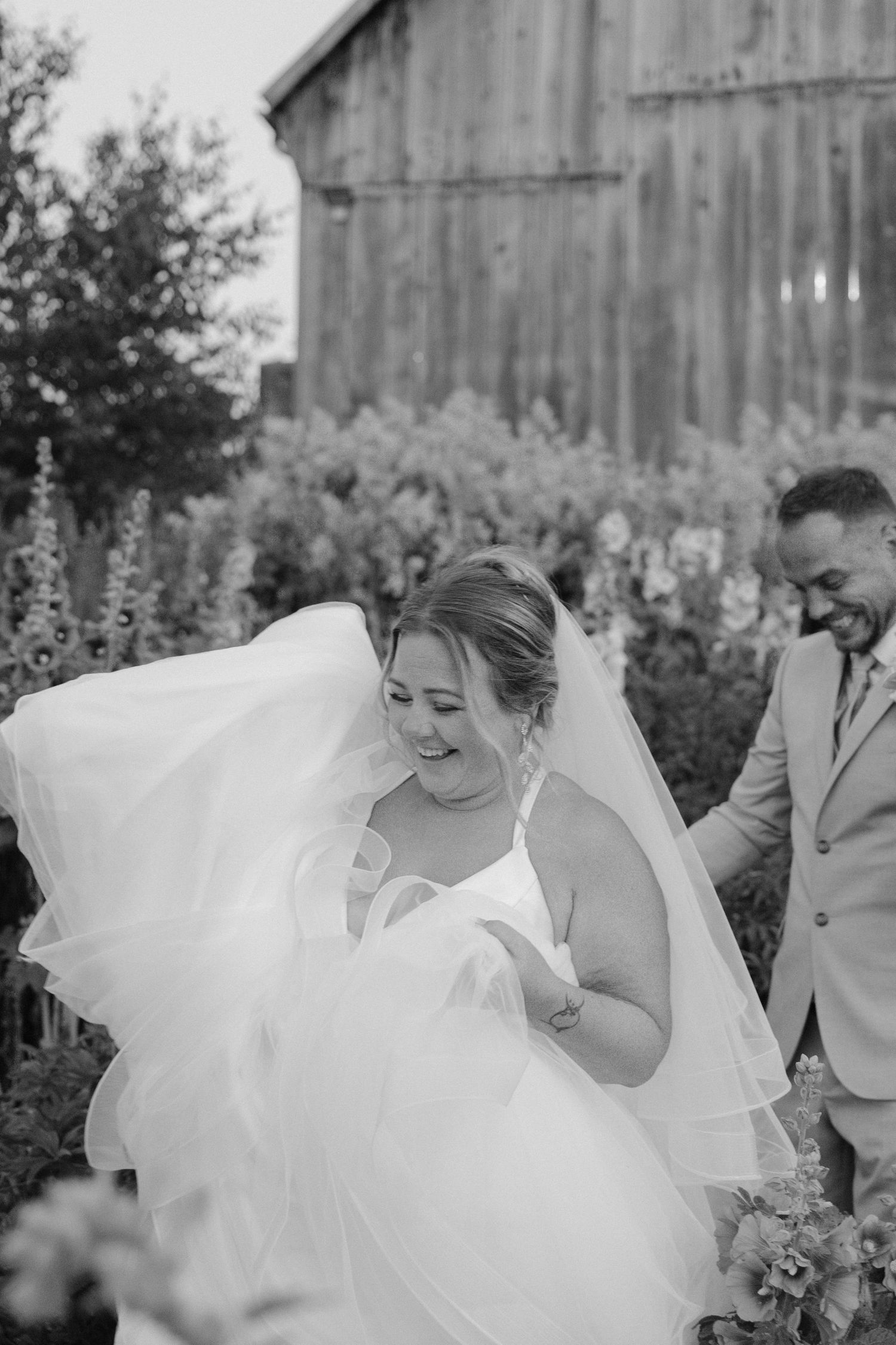 Black and white photo of laughing bride with flowing veil caught in breeze beside groom in barn garden.