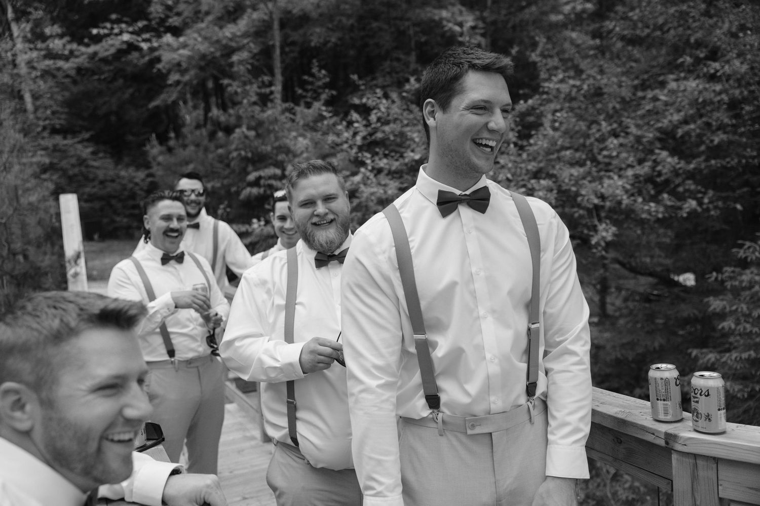 Black and white candid photo of laughing groomsmen in white shirts and suspenders outdoors.