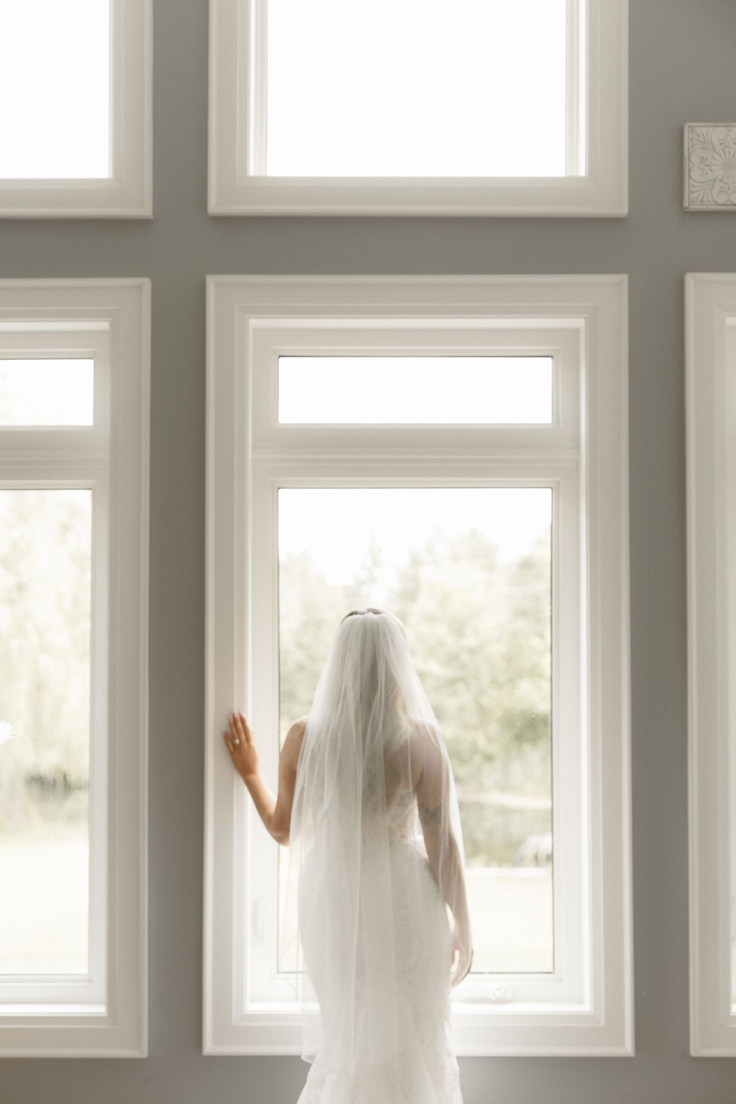 Bride in white gown and veil standing before large windows in soft natural light.