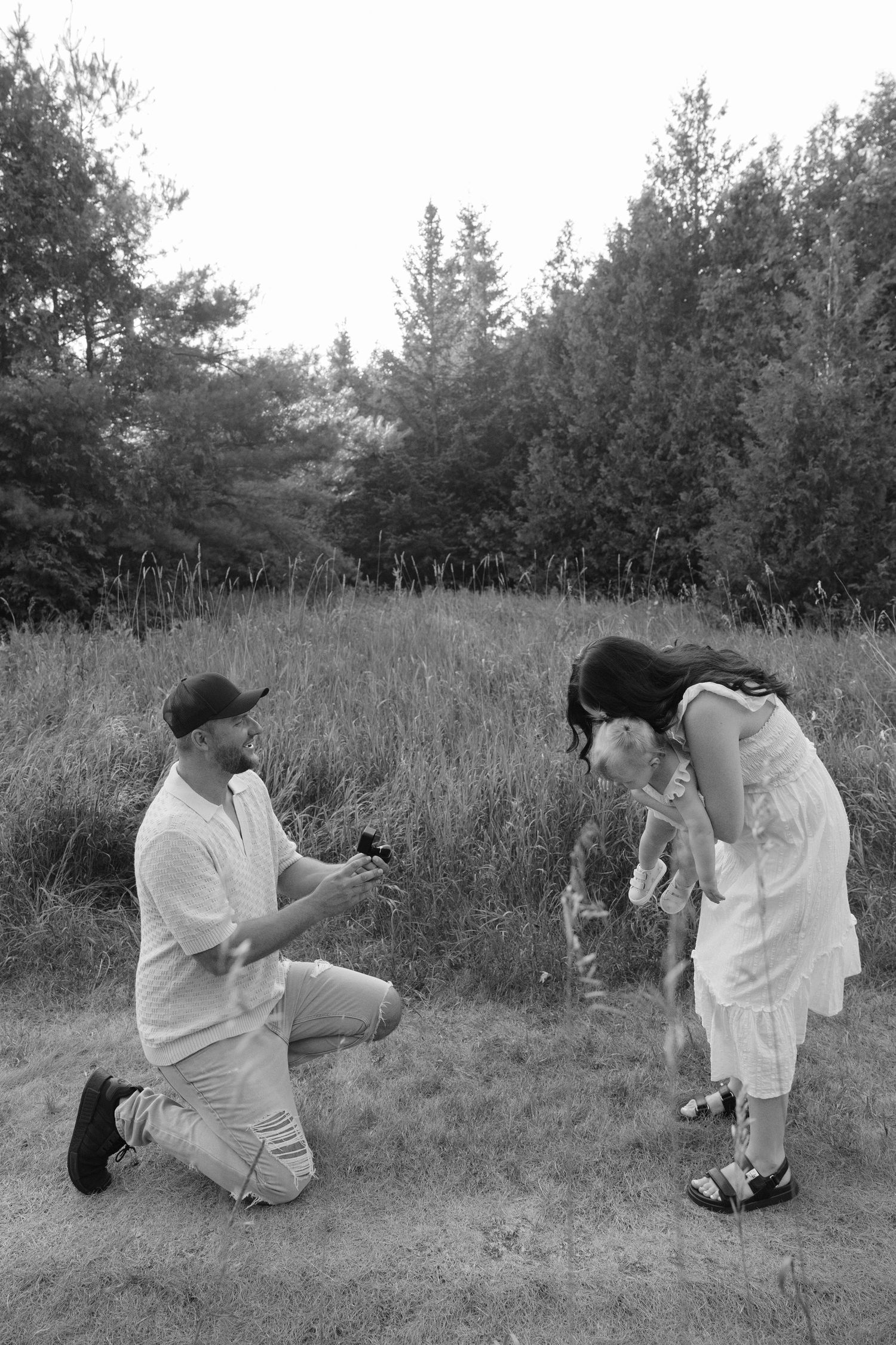 Black and white photo of a man proposing on one knee to a woman and child.