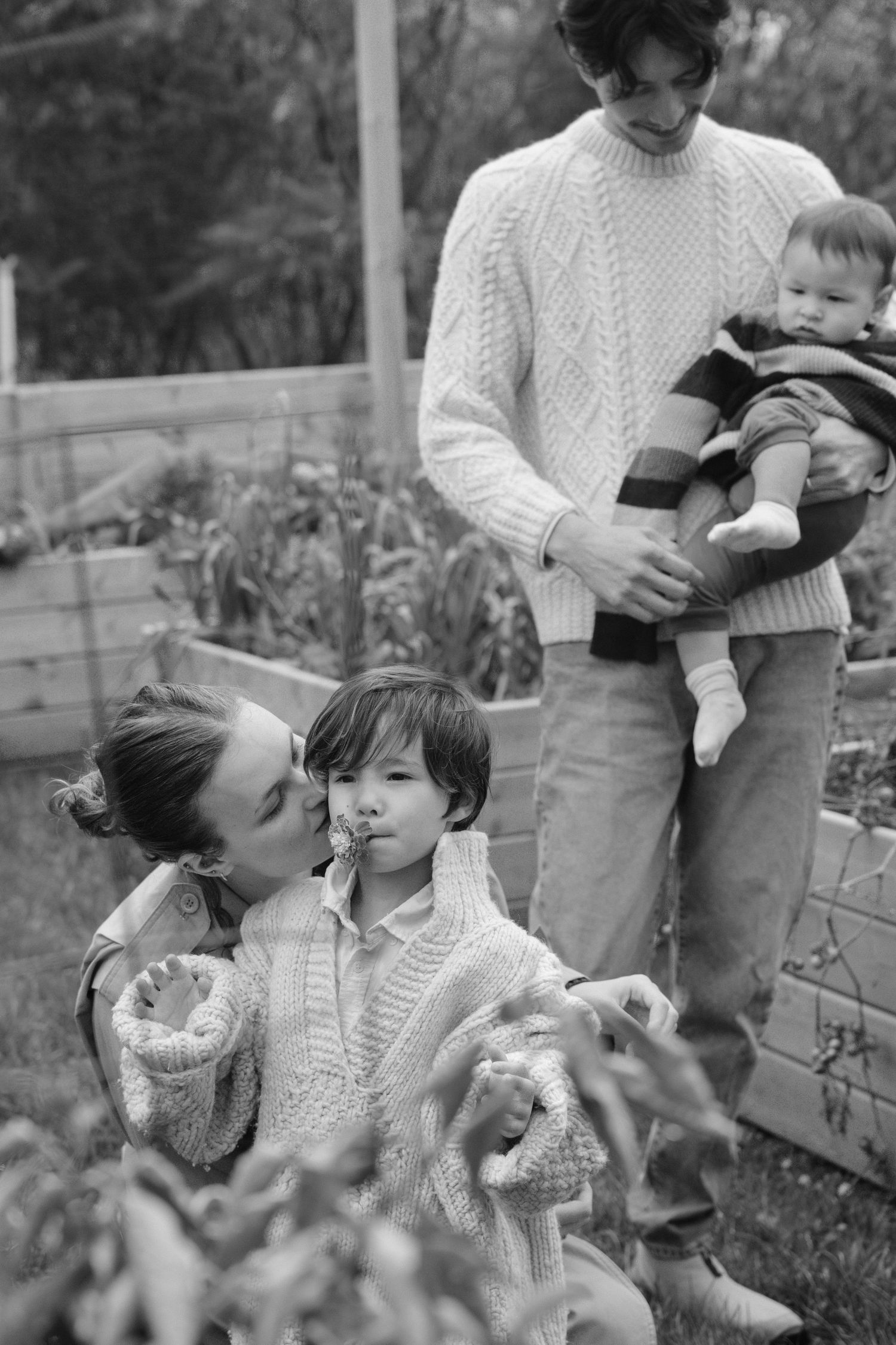 Black and white family photo with mother kissing child outdoors in garden.