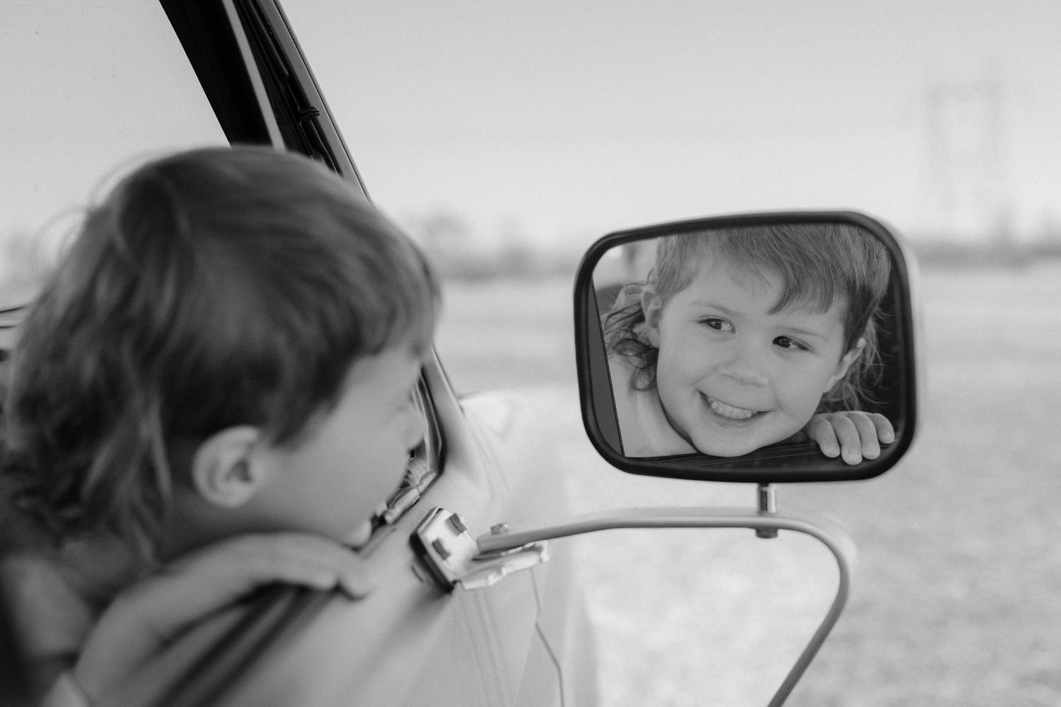 Black and white photo of a child leaning out a car window smiling at their reflection in the mirror.
