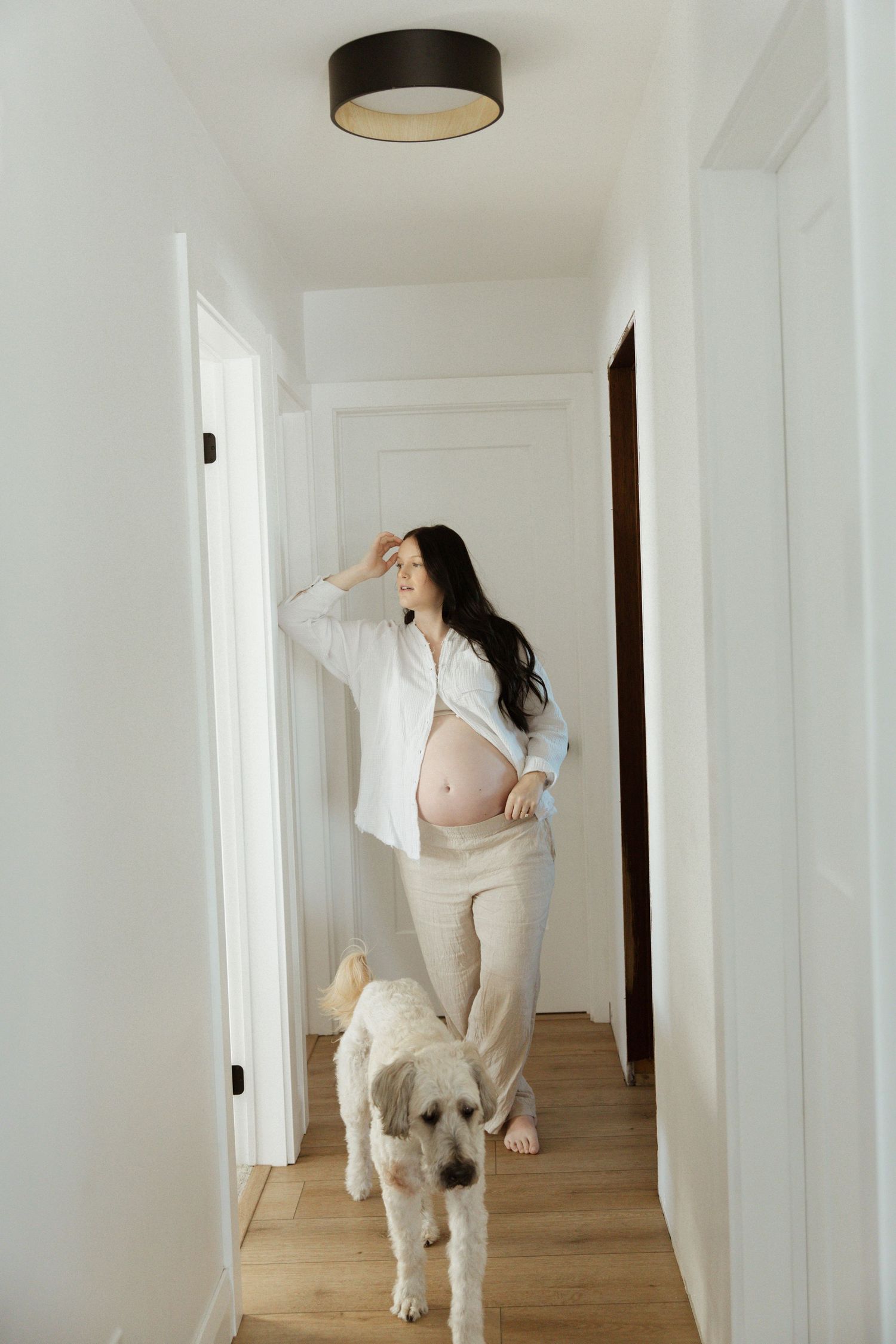 Pregnant woman in white shirt stands in bright hallway with fluffy dog, cradling baby bump.