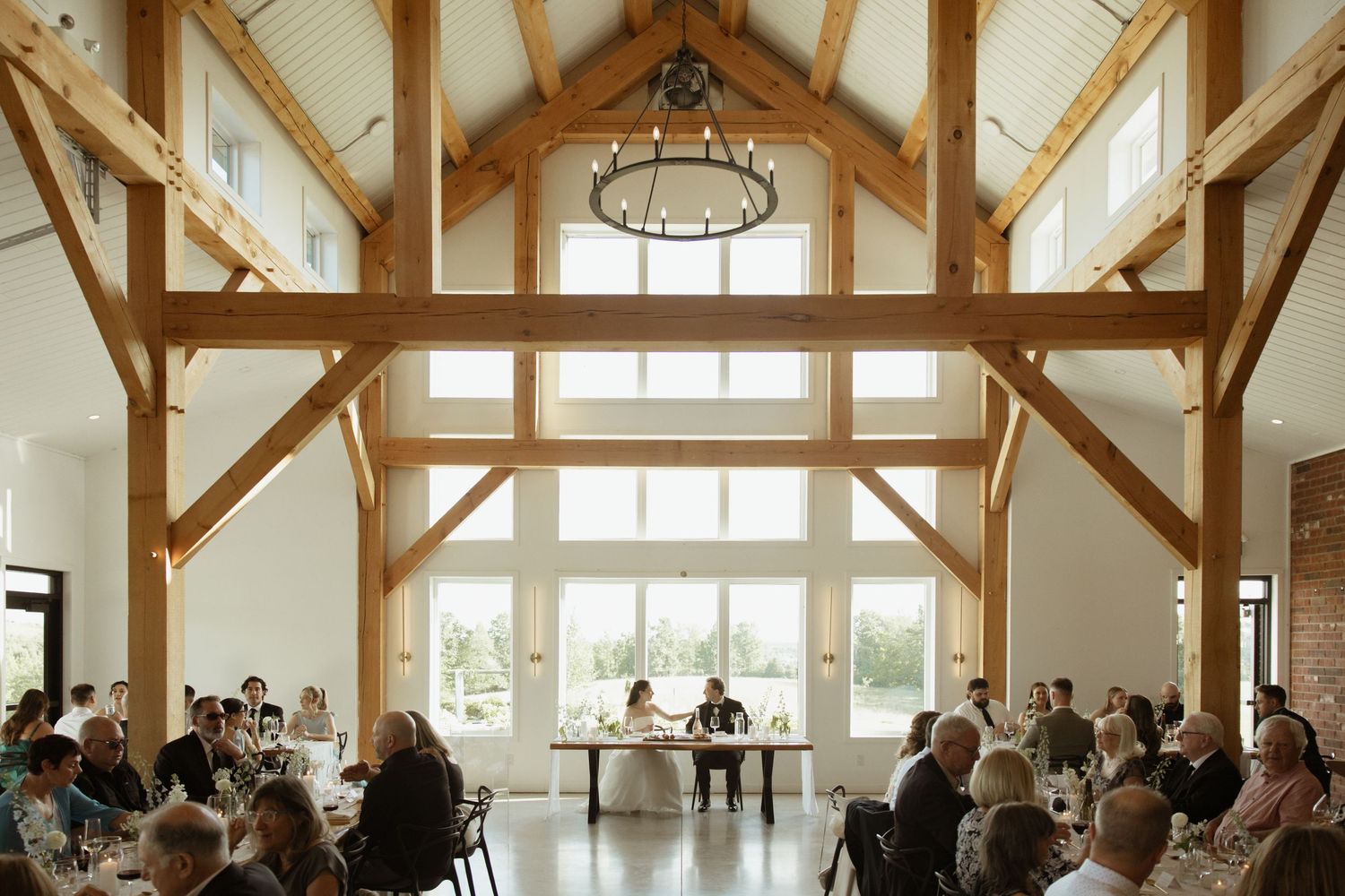Elegant barn wedding reception with soaring timber frame ceilings, chandelier, and guests seated at round tables.