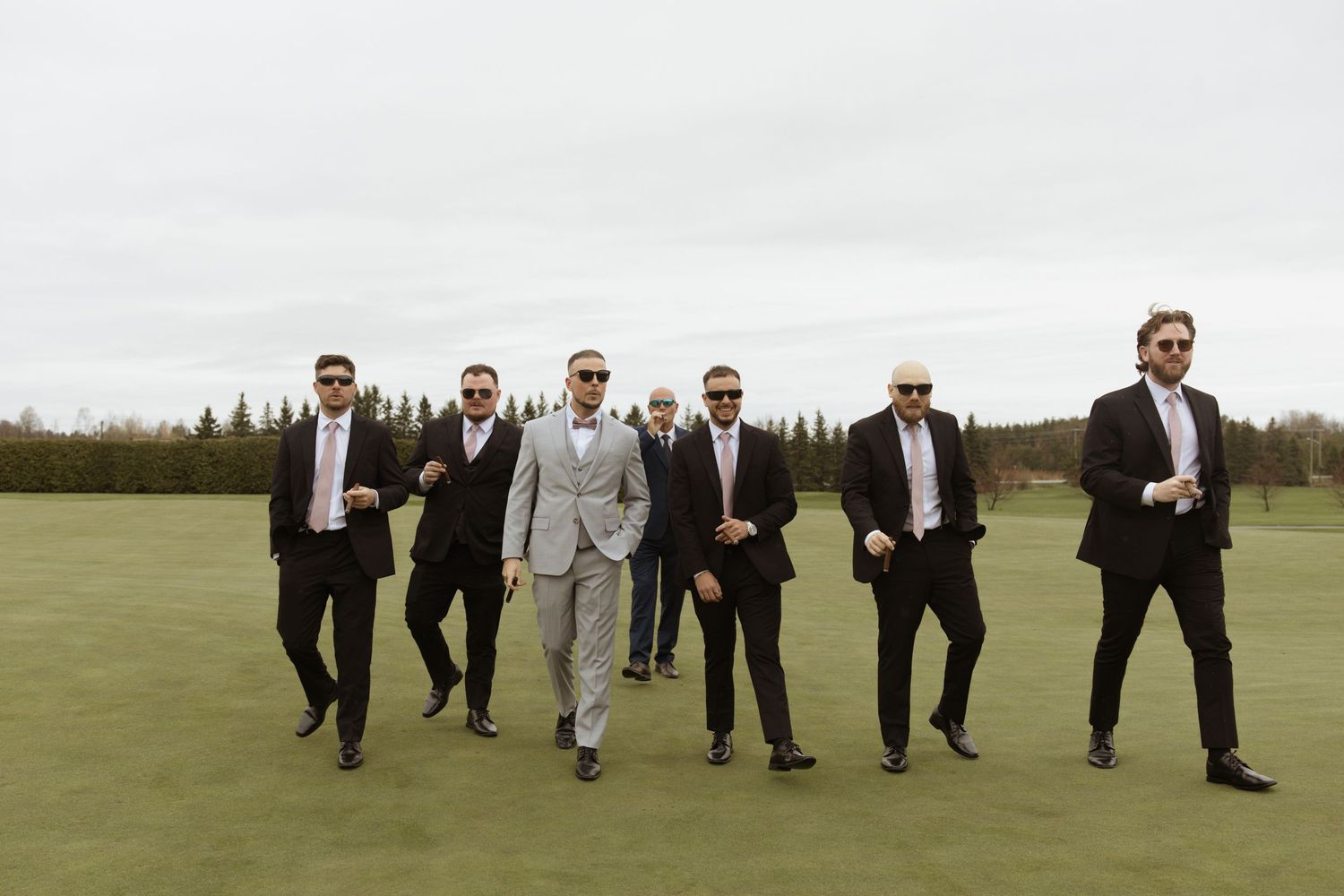 Groom and groomsmen in suits and sunglasses walking confidently on a golf course green at an outdoor wedding.