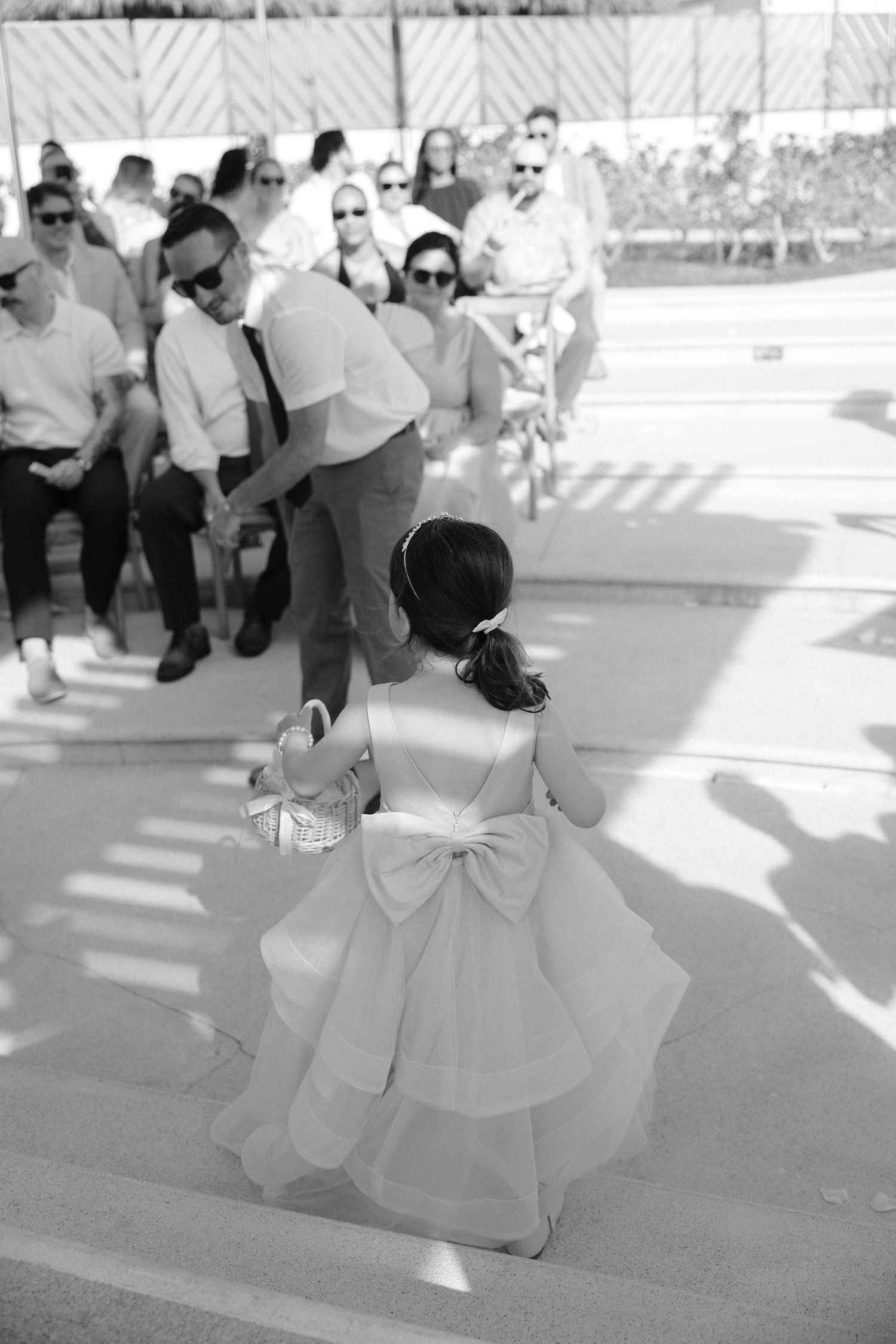 Flower girl in white dress with bow walks down aisle escorted by man at outdoor wedding ceremony.