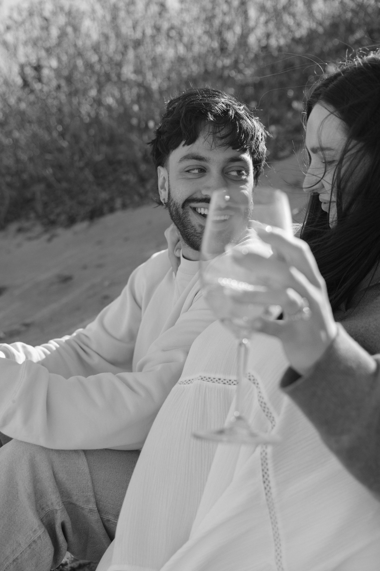 Black and white photo of smiling groom in white kurta raising champagne glass with bride outdoors on sandy ground.