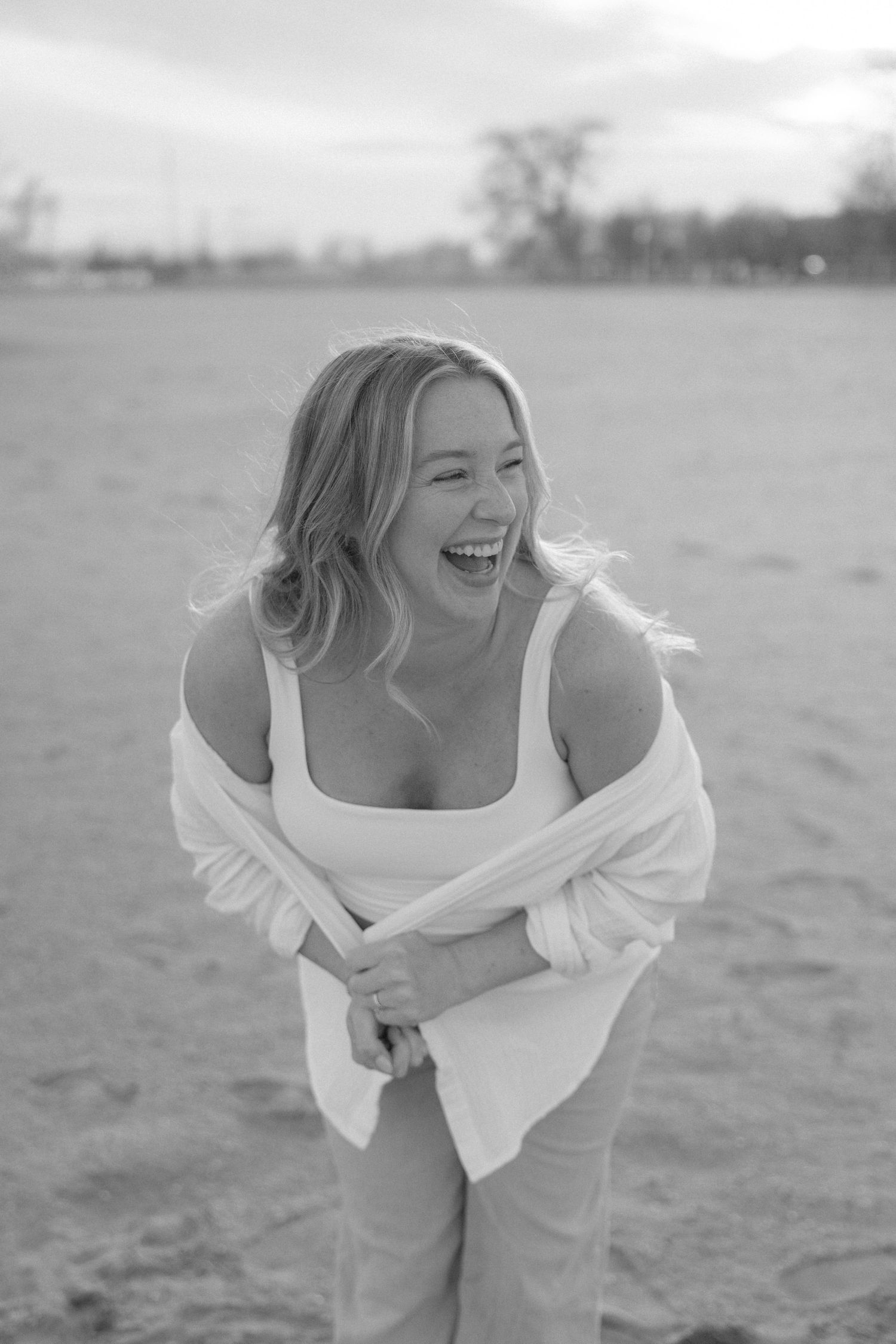 Black and white photo of a laughing blonde woman in a white outfit enjoying a fun moment on a beach.
