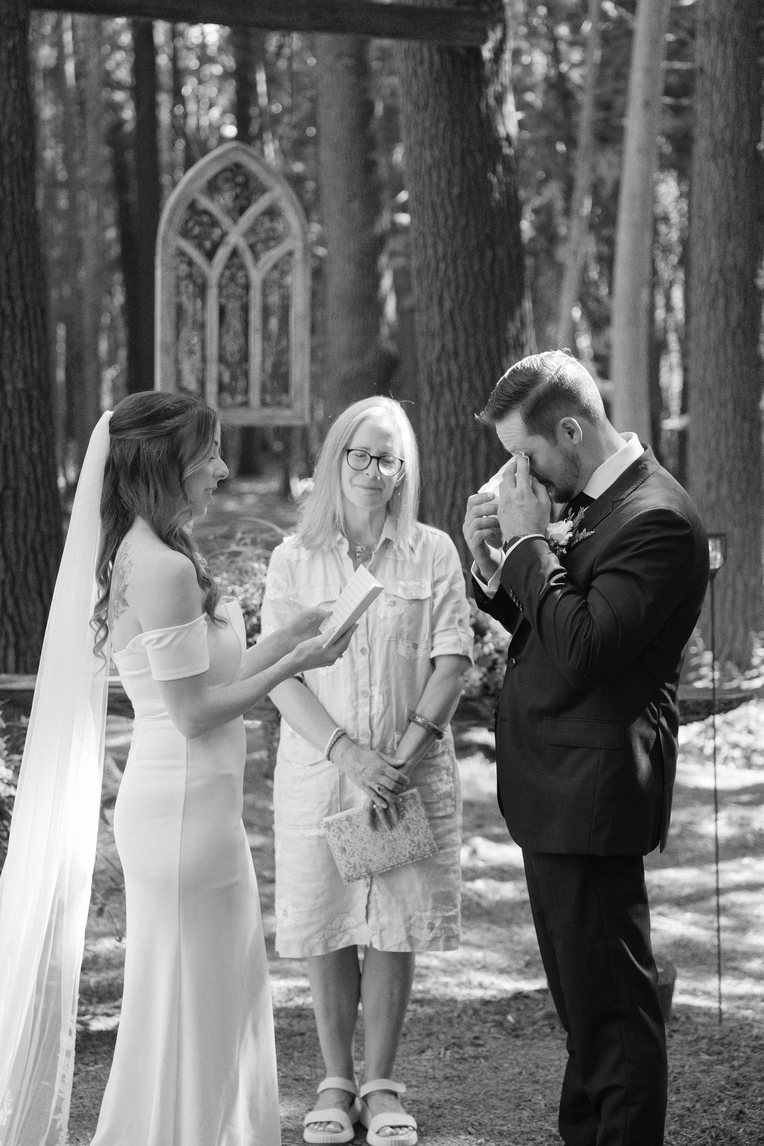Emotional black and white photo of groom wiping tears during outdoor forest wedding vow exchange.