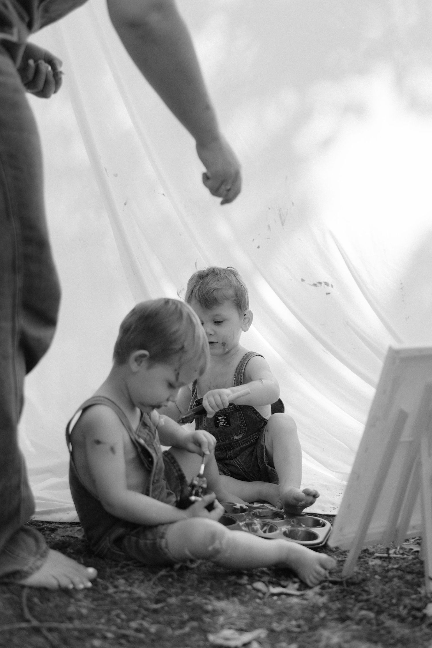 Black and white image of two toddlers in overalls sitting on ground with paint palette near white backdrop outdoors.