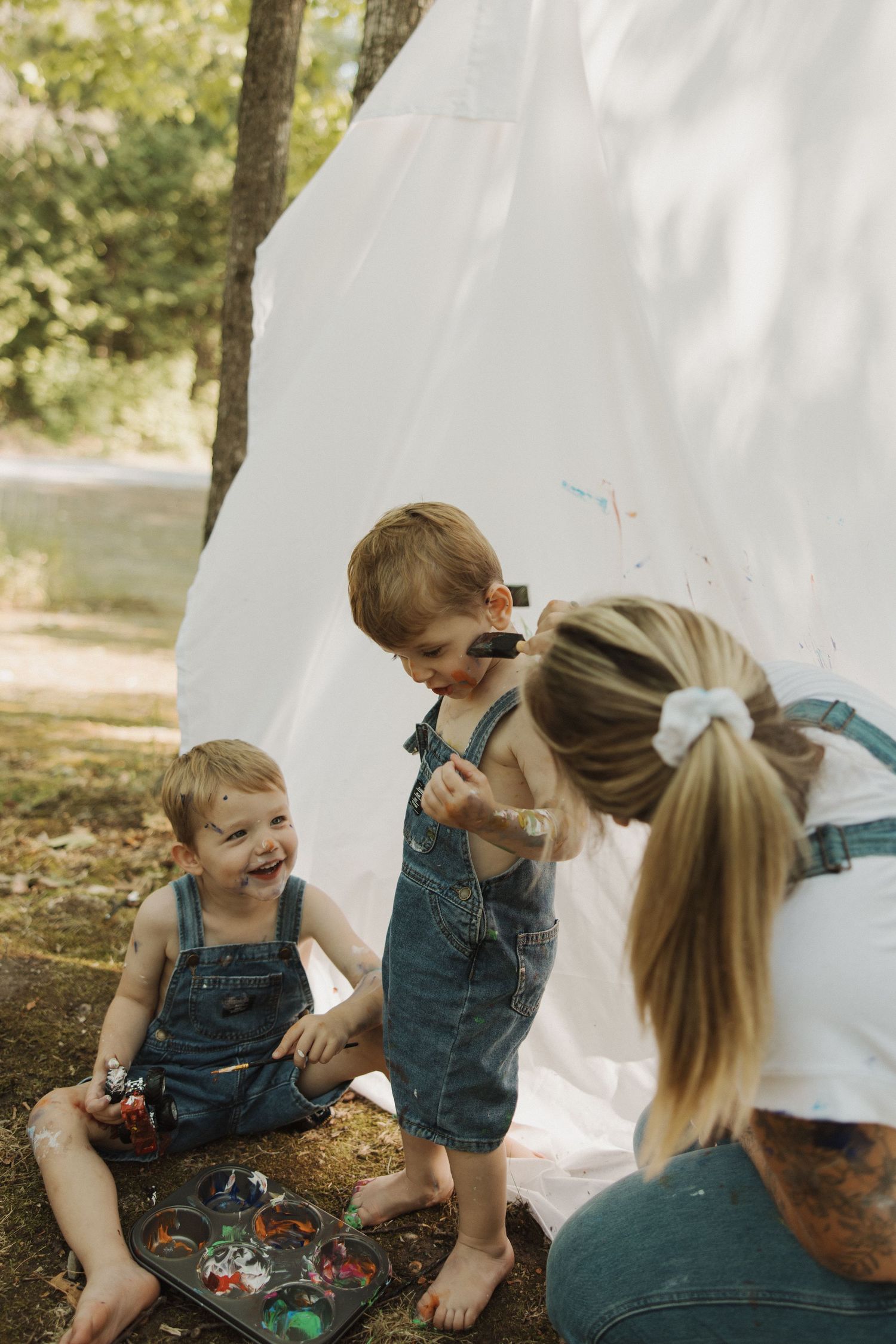 Mother with hair bow interacting with two young boys in denim overalls doing art near white canvas in forest.