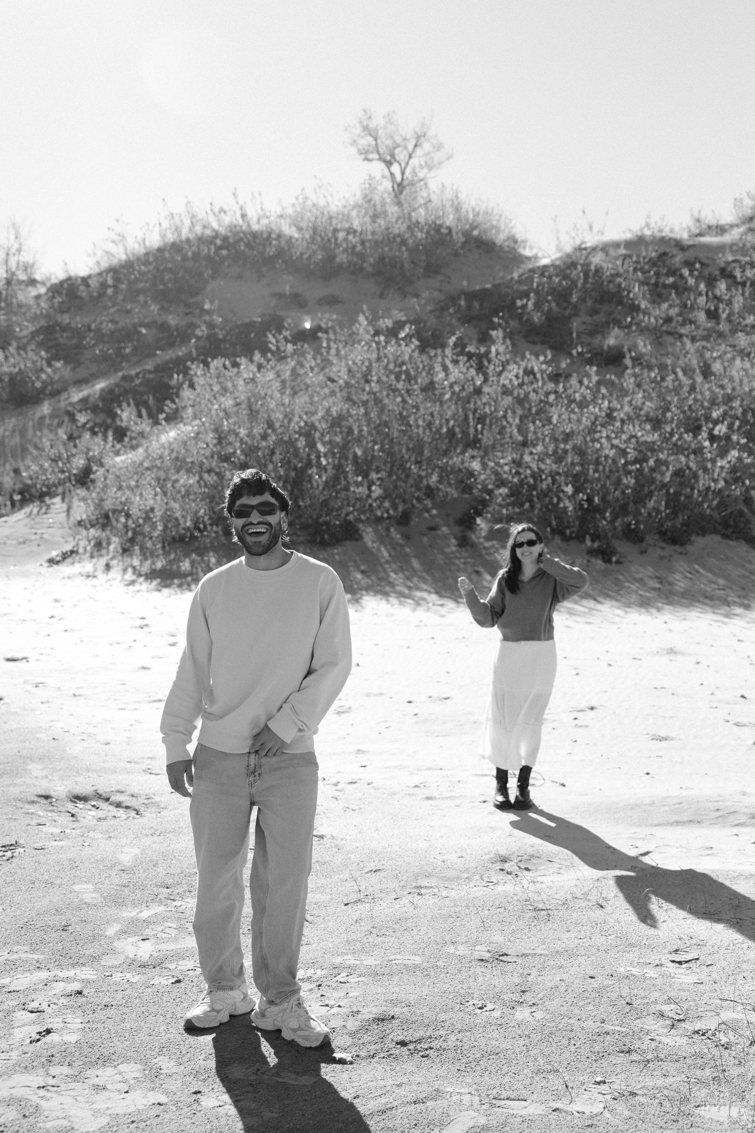 Couple posing in black and white on a sunny sandy landscape with desert shrubs in background.