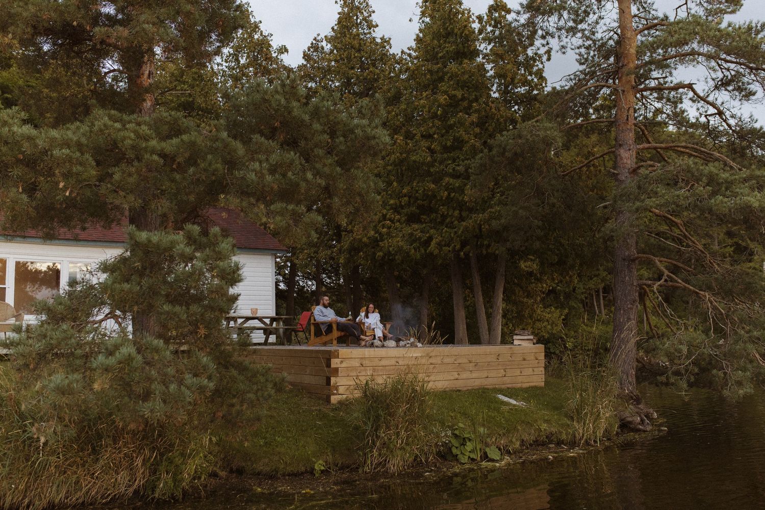 A cozy lakeside cottage with a wooden deck nestled among tall pine trees on a calm waterway.