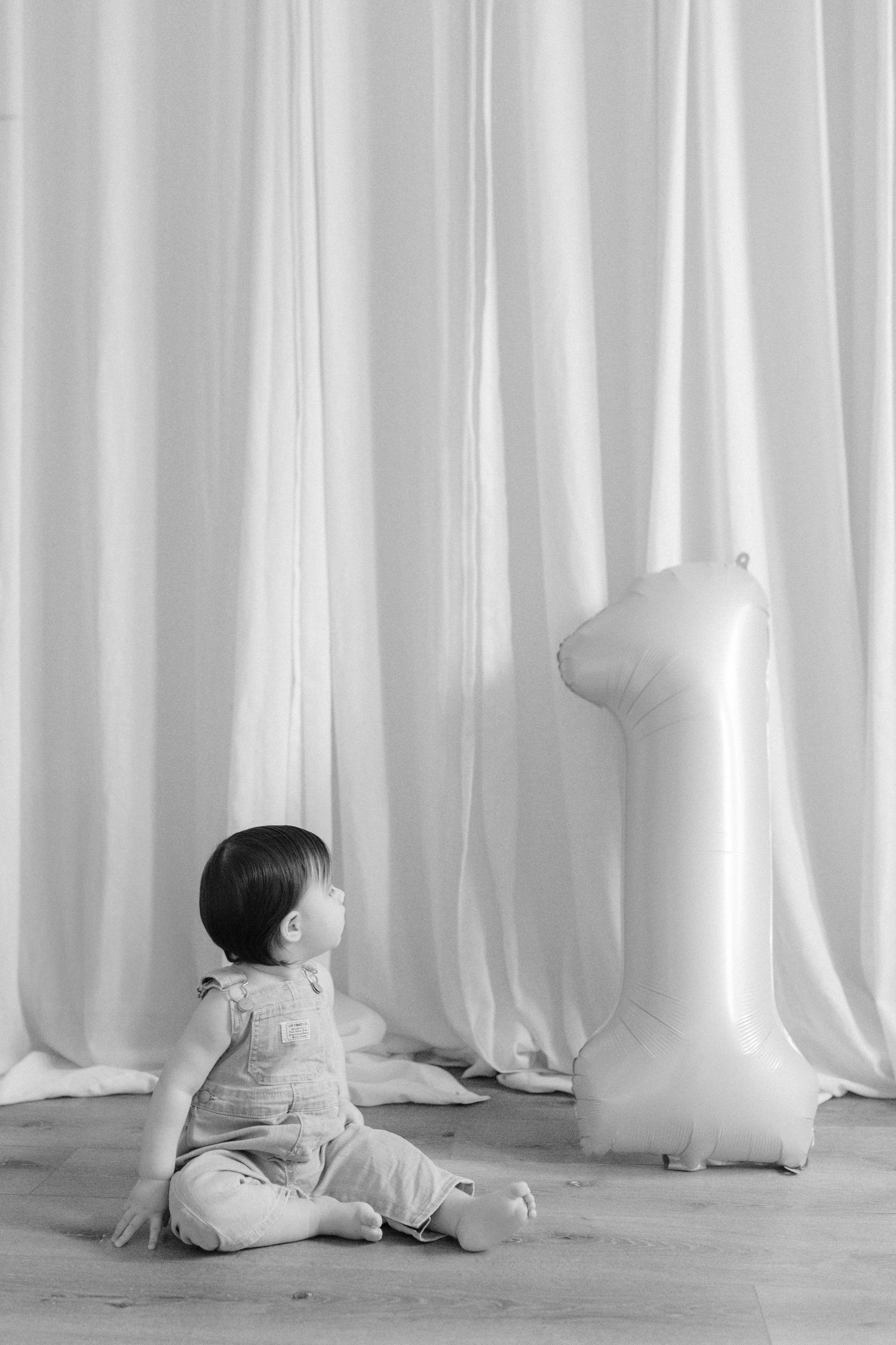 Black and white photo of a baby sitting on the floor next to a number one balloon in front of white curtains.