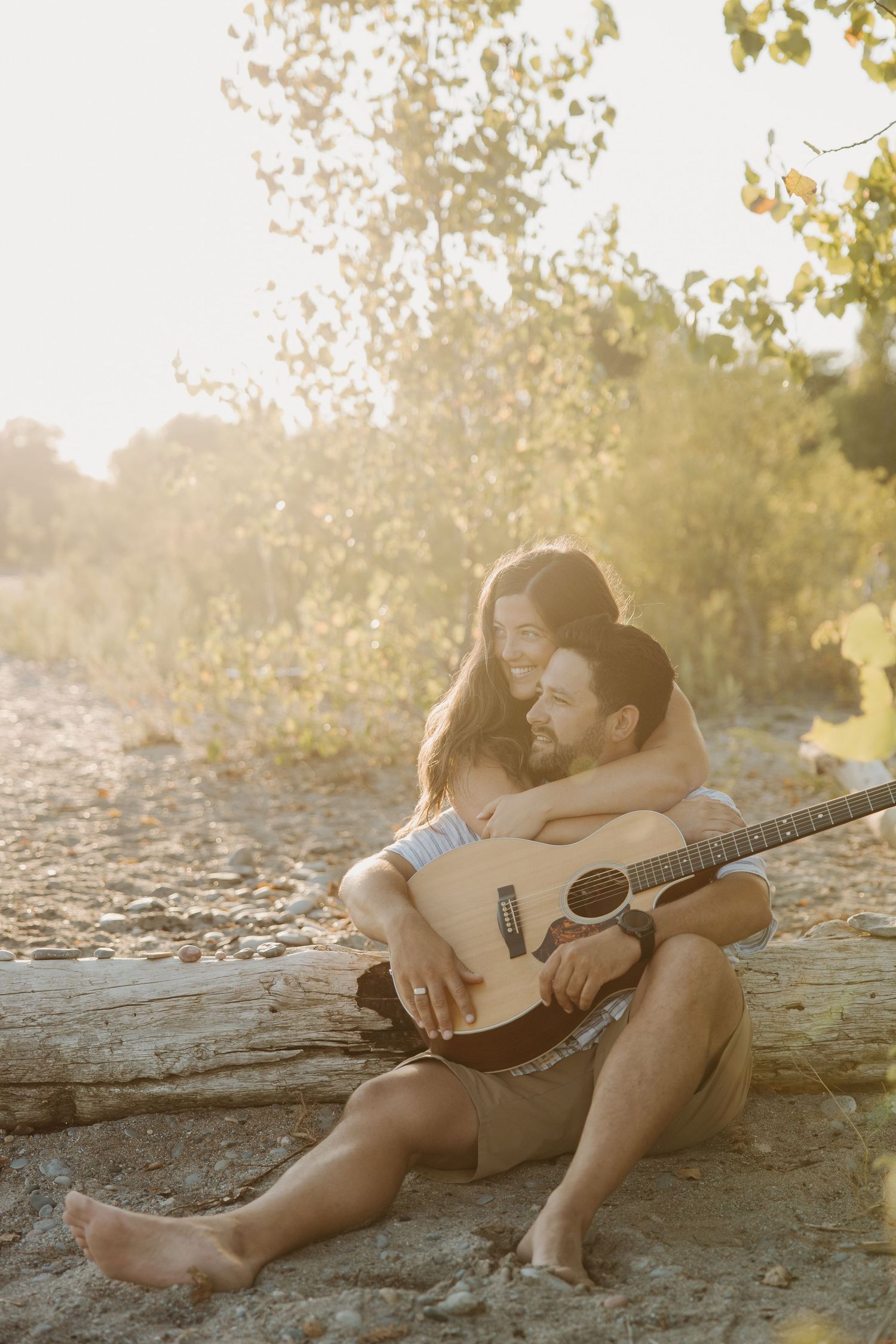 Couple sitting on a log at golden hour, woman embracing man from behind as he holds an acoustic guitar.