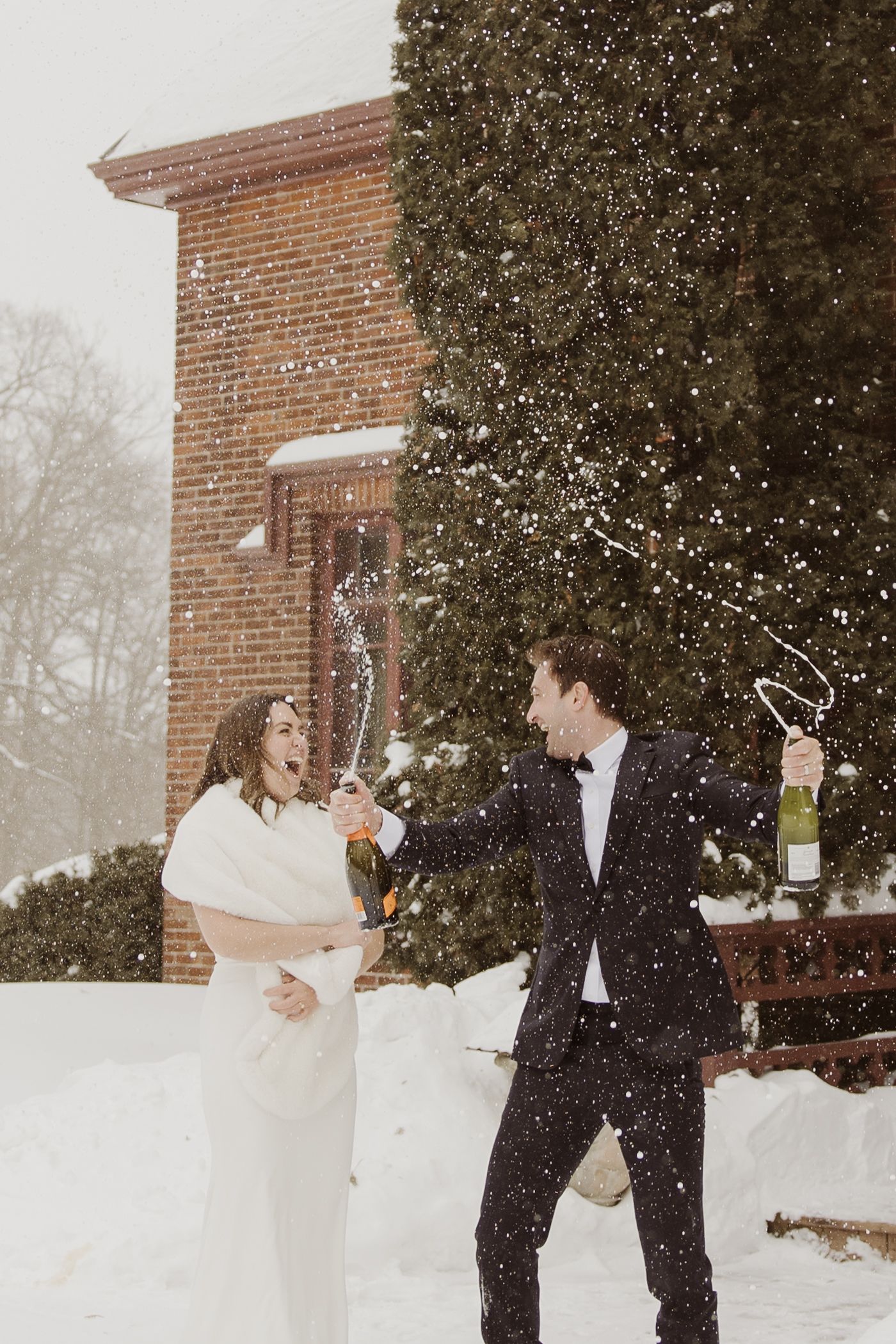 Bride and groom popping champagne in the snow outside a brick building during a winter wedding celebration.