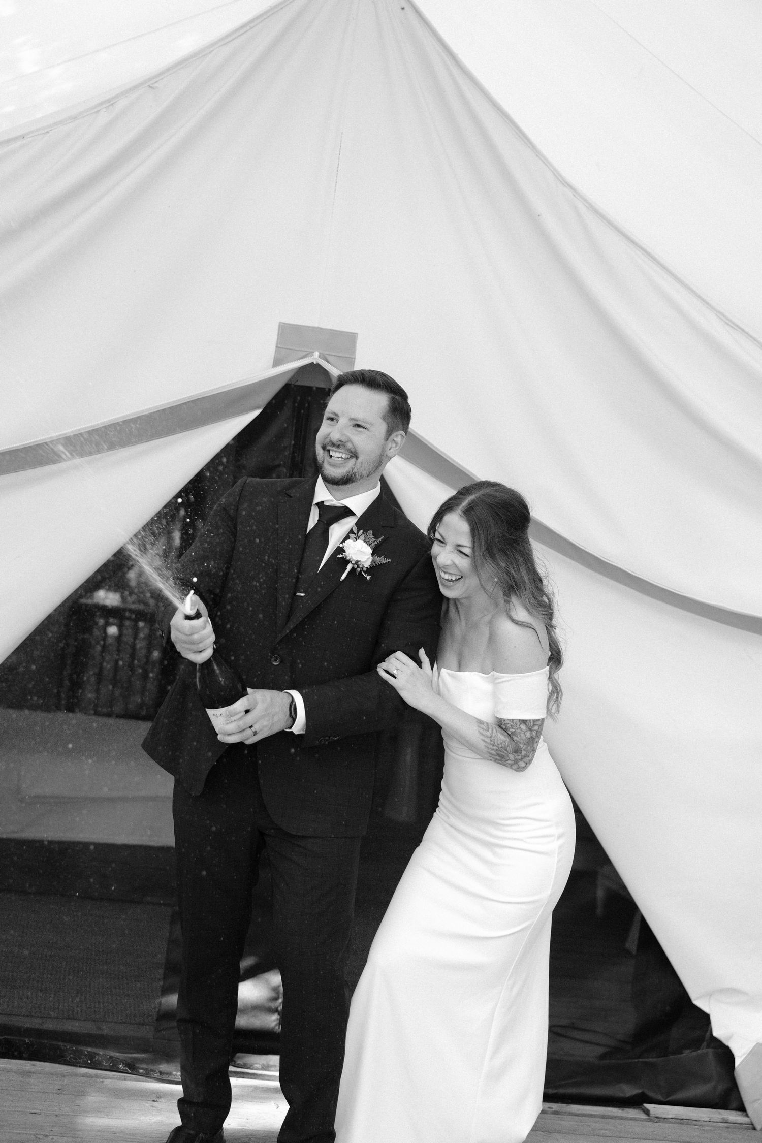 Black and white photo of a laughing bride and groom popping champagne under a white tent at their wedding reception.