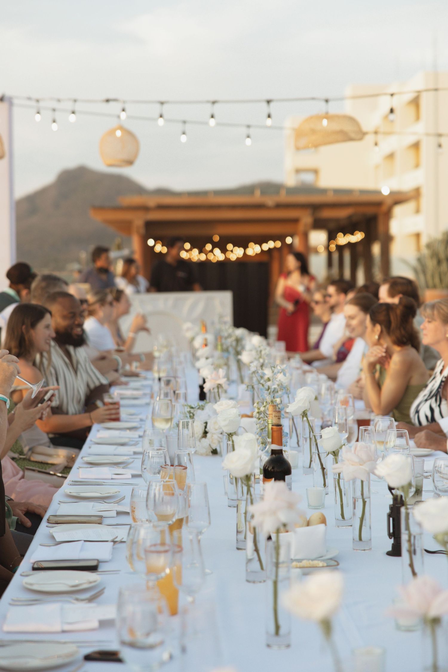 Guests seated at a long elegant outdoor wedding reception table adorned with white roses and string lights at dusk.