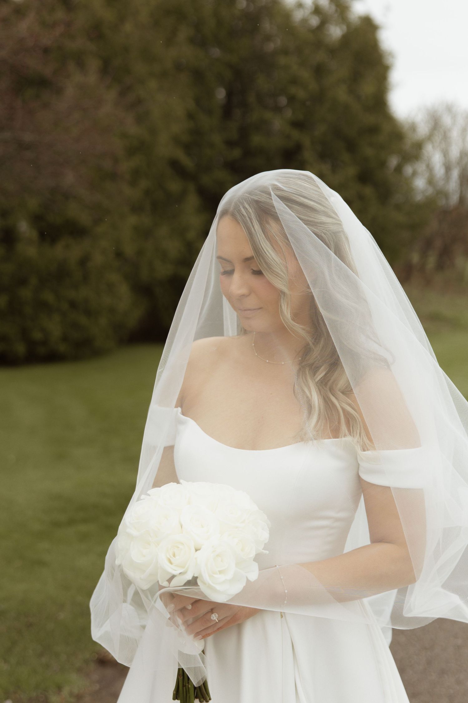 Bride in white off-shoulder gown and veil holds white rose bouquet outdoors, looking down gracefully.