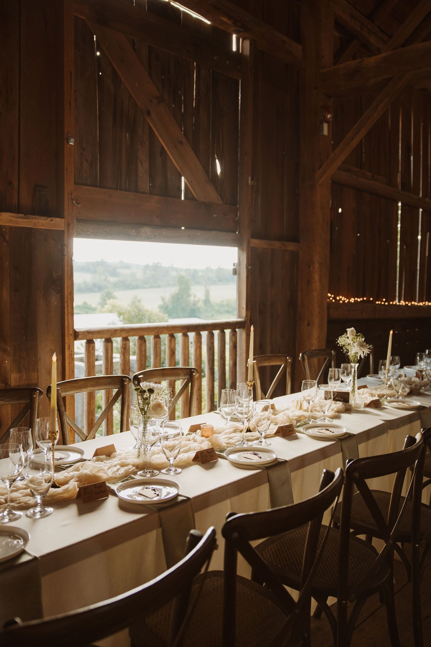 Elegant barn wedding reception table set with candles, glassware, and floral centerpieces with scenic countryside view.