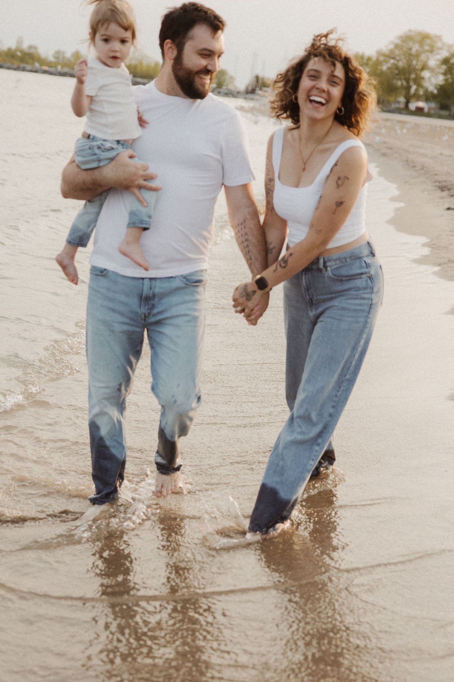 A happy couple in white shirts and jeans walks along the beach holding their toddler.