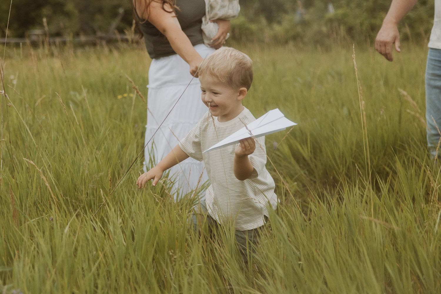 A young child runs through tall grass holding a paper airplane with parents nearby.
