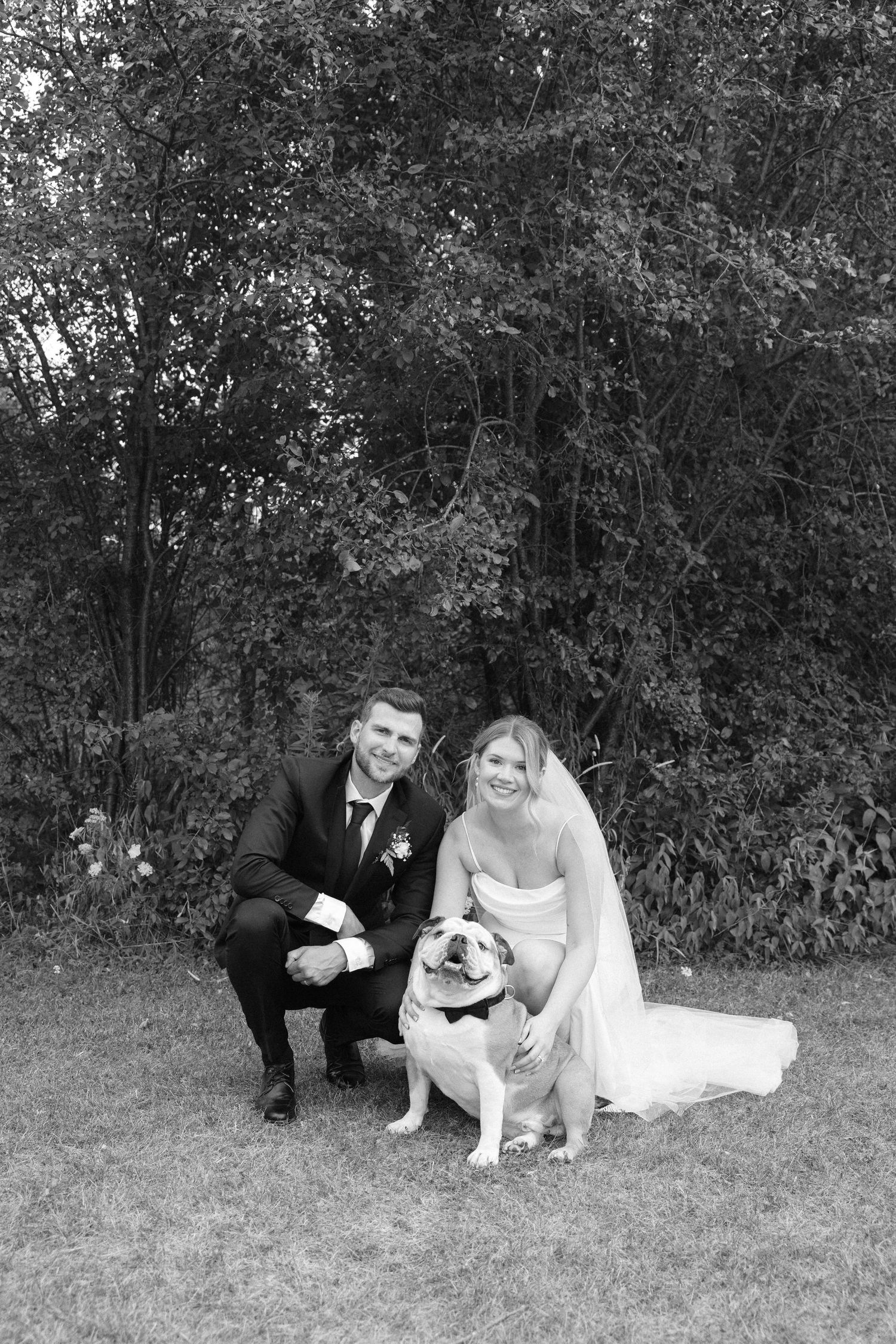 Black and white wedding portrait of bride and groom posing with their English Bulldog outdoors.