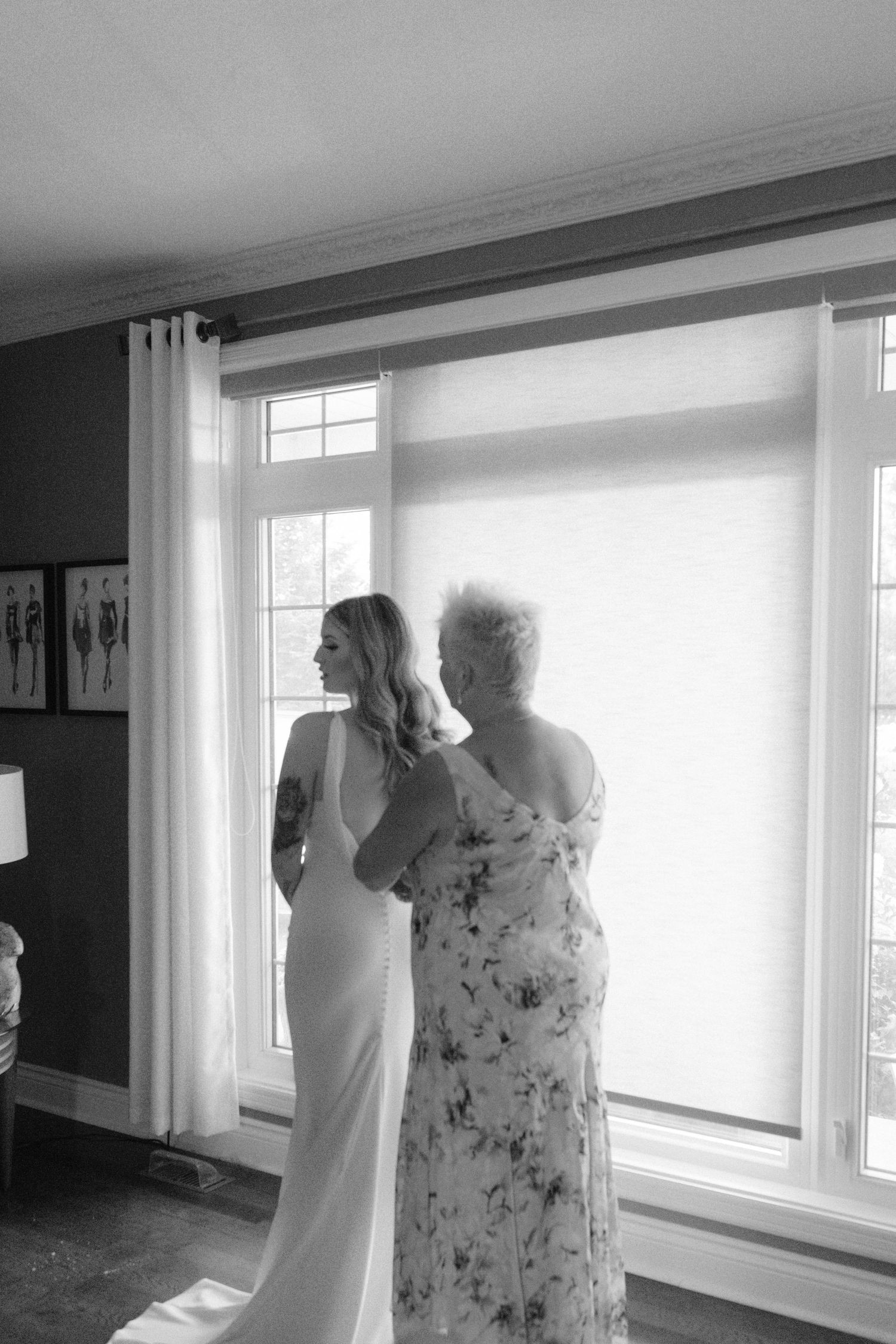 Black and white photo of bride in elegant gown with older woman helping her prepare by a bright window.
