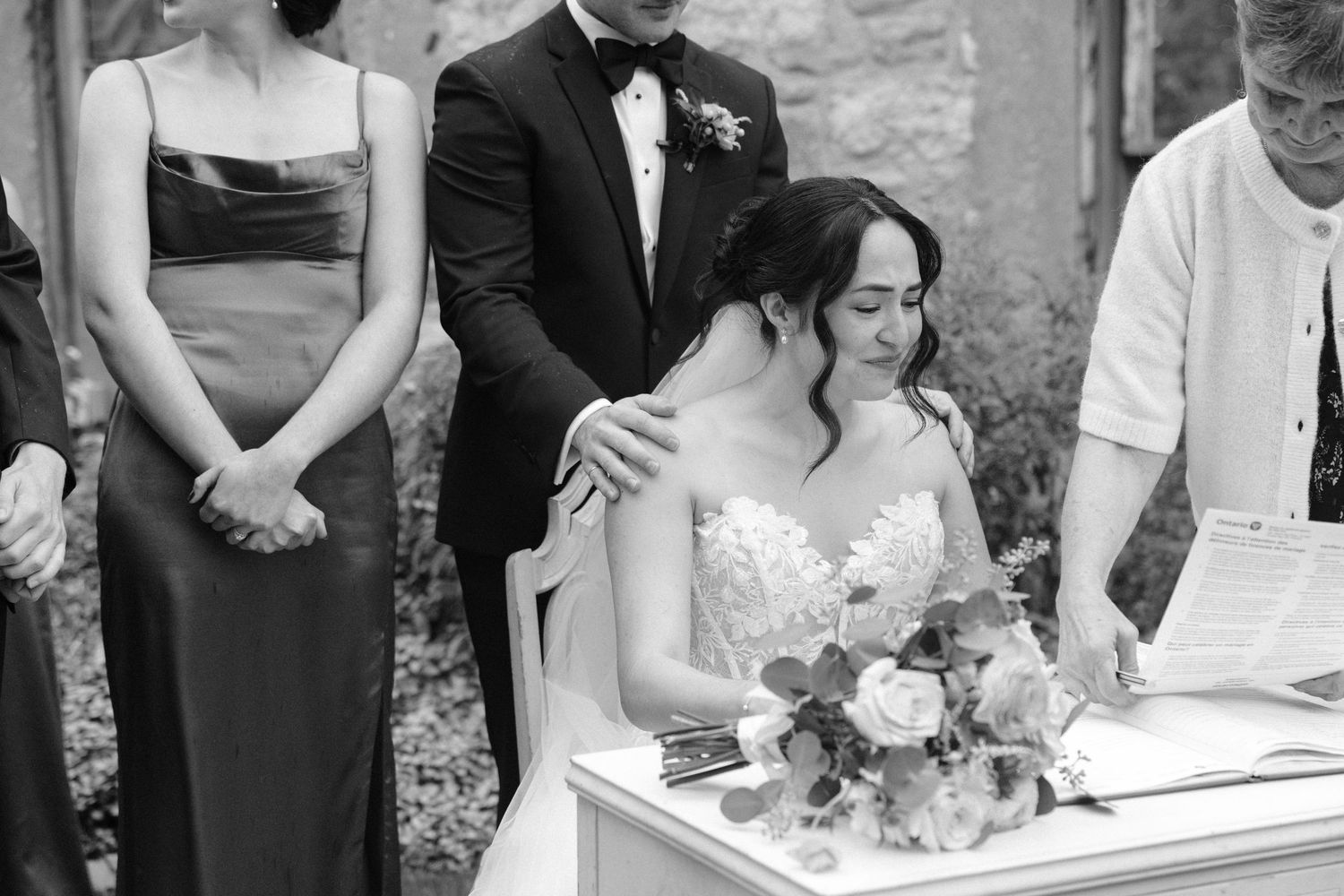 Black and white photo of emotional bride signing marriage certificate with groom comforting her.