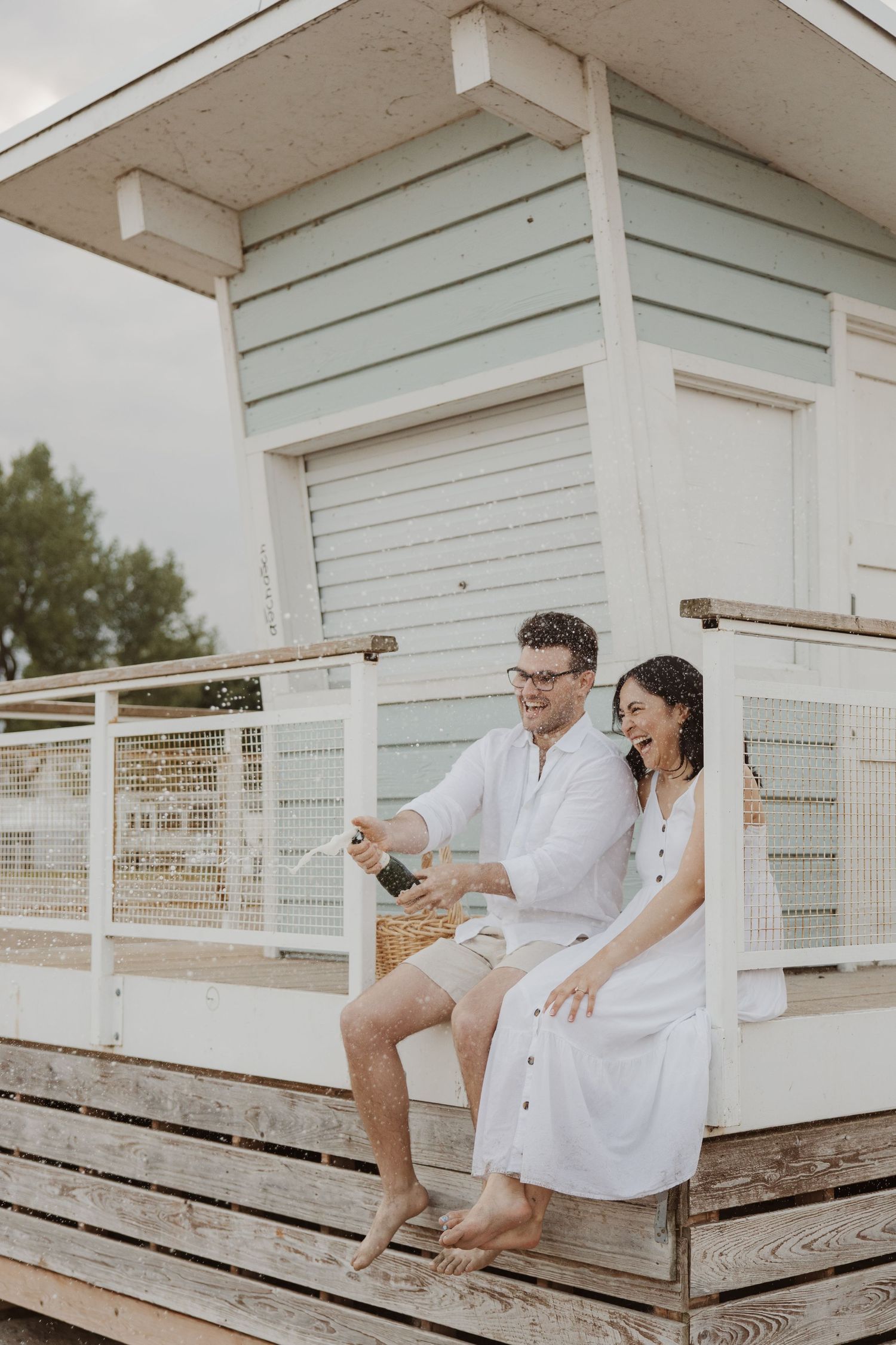 Couple popping champagne bottle while sitting on beach hut porch together.