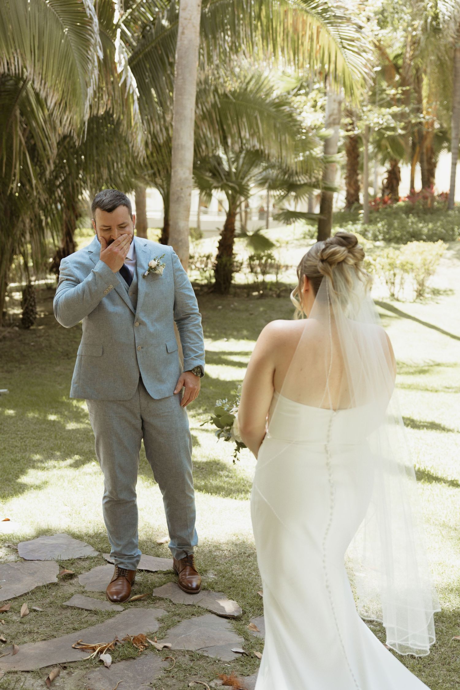 Groom in light blue suit covers mouth emotionally as bride in white gown approaches for first look outdoors.
