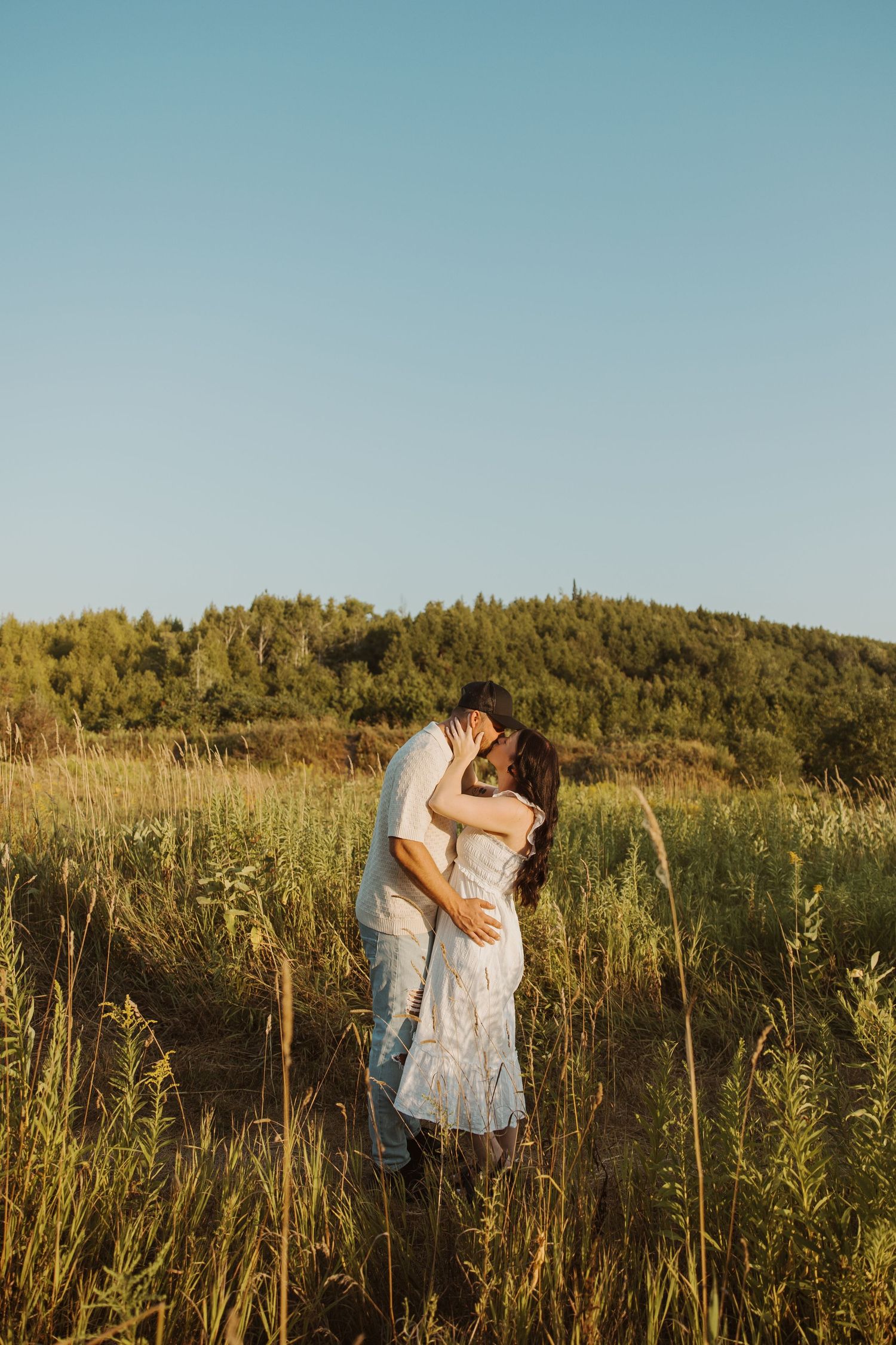 Couple sharing a kiss in a golden field of tall grass with a forested hill and blue sky behind them.