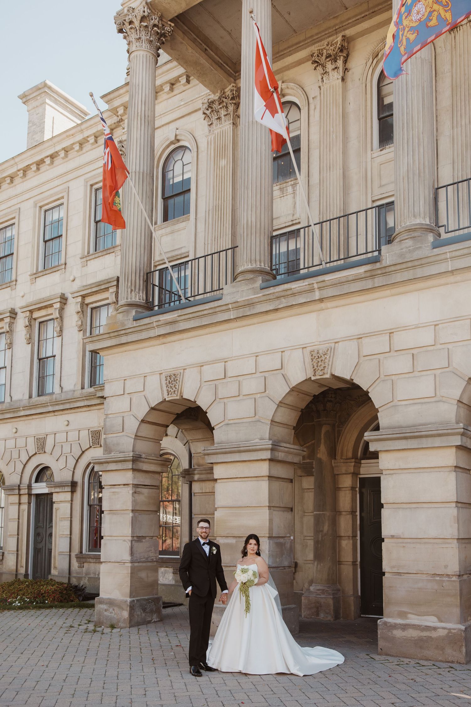 Bride in a full white gown and groom in a black tuxedo posing in front of a historic building with flags.