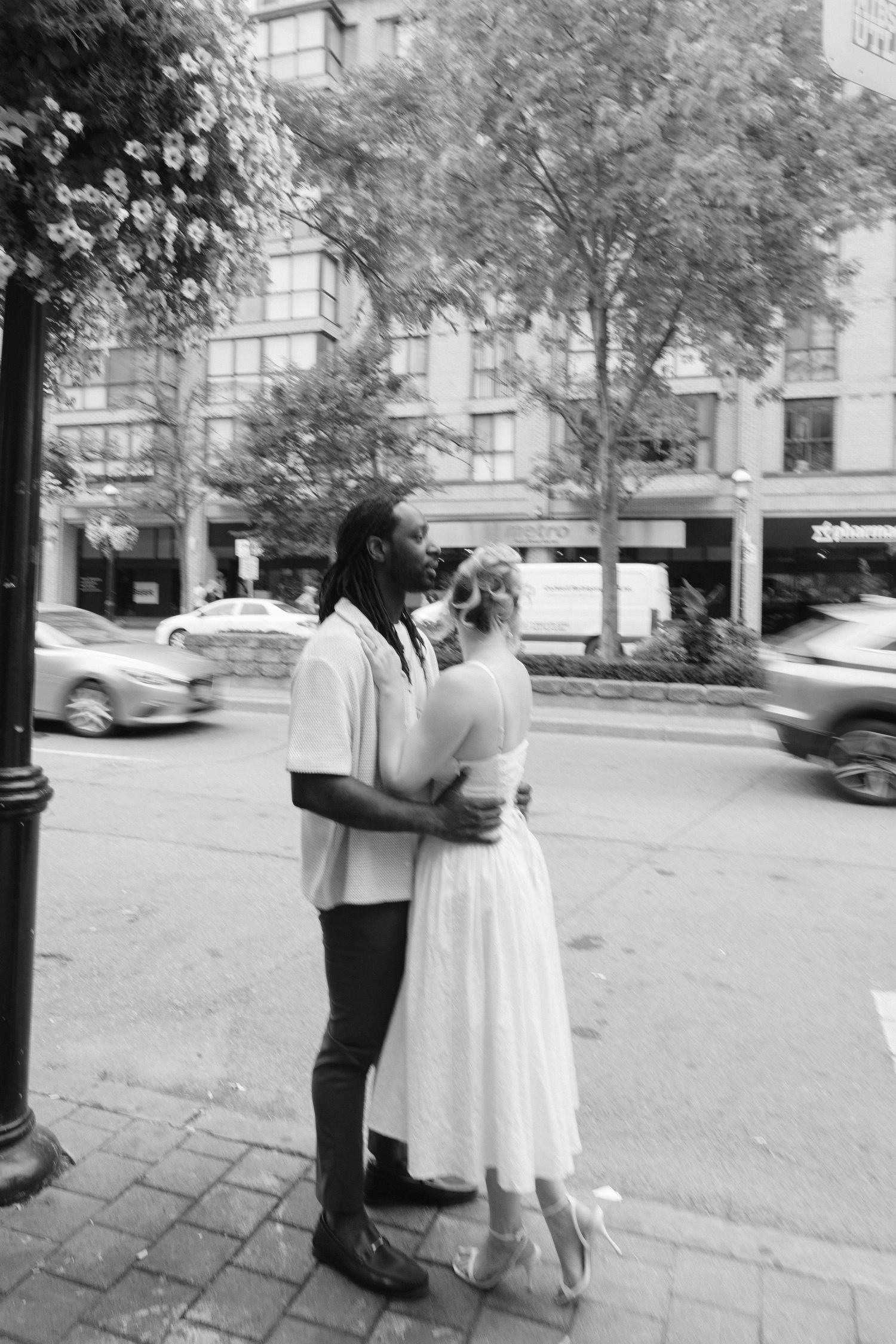 Black and white photo of interracial couple embracing on a city street corner surrounded by urban scenery.
