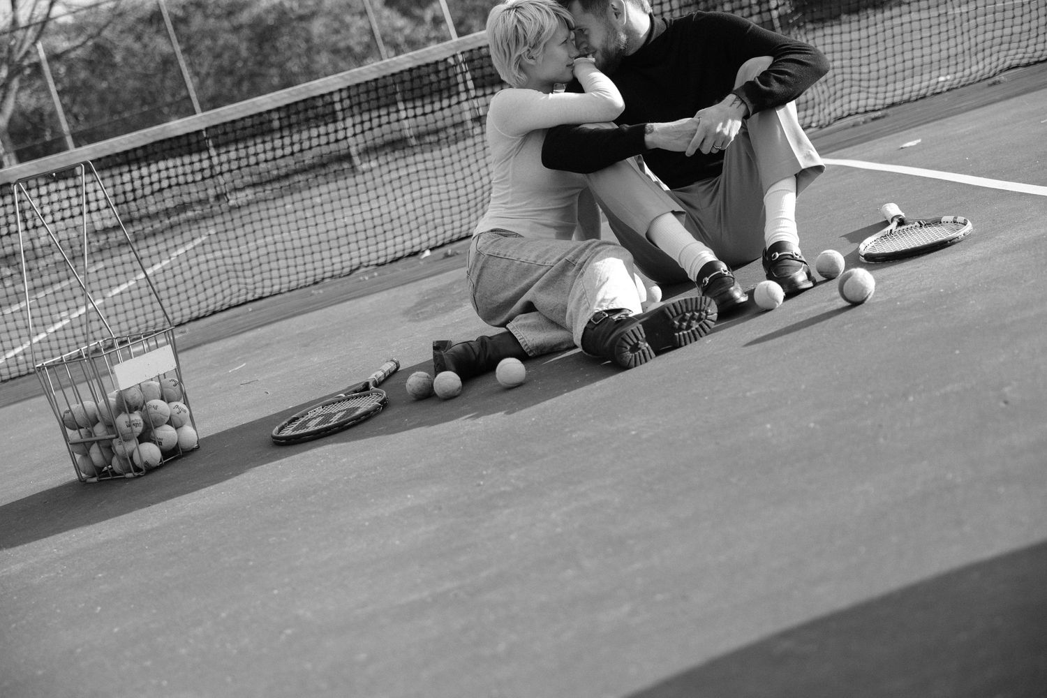 Black and white photo of couple sitting on tennis court surrounded by balls and rackets.