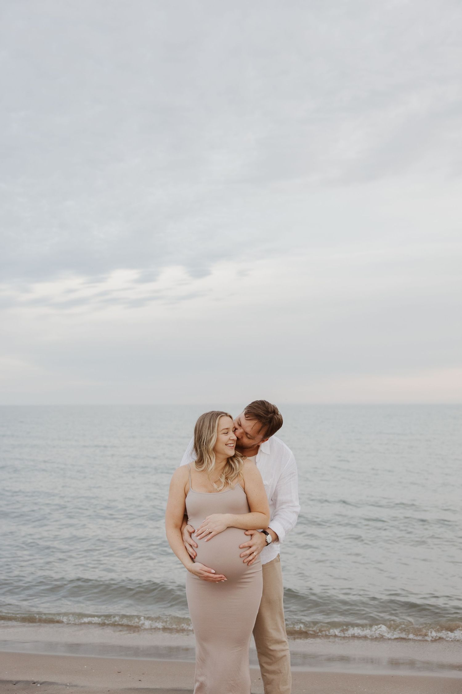 Couple sharing a tender moment on a beach, man kissing pregnant woman's cheek while holding her bump.