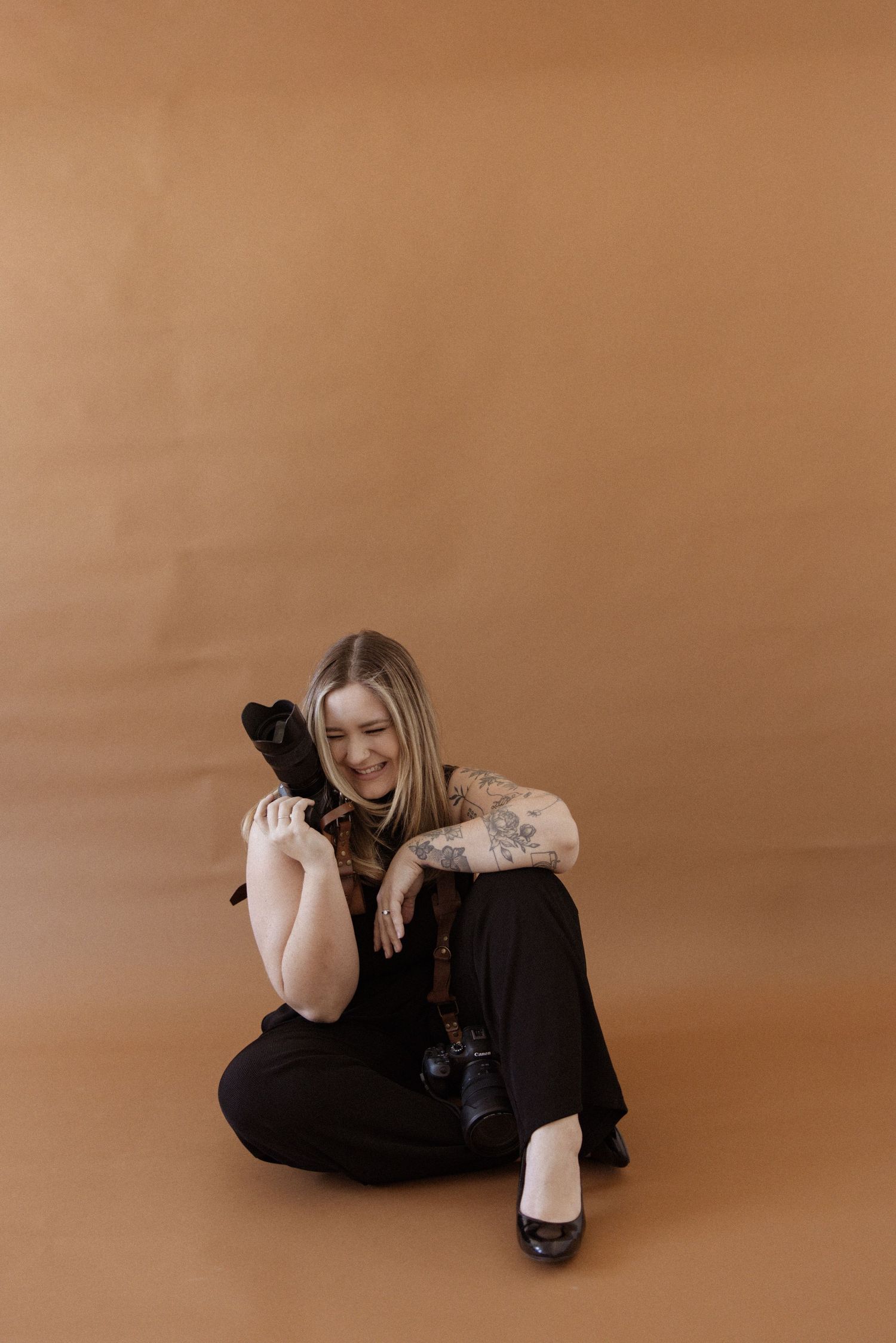 Smiling tattooed blonde woman sitting on floor hugging camera, wearing black outfit against warm tan studio background.