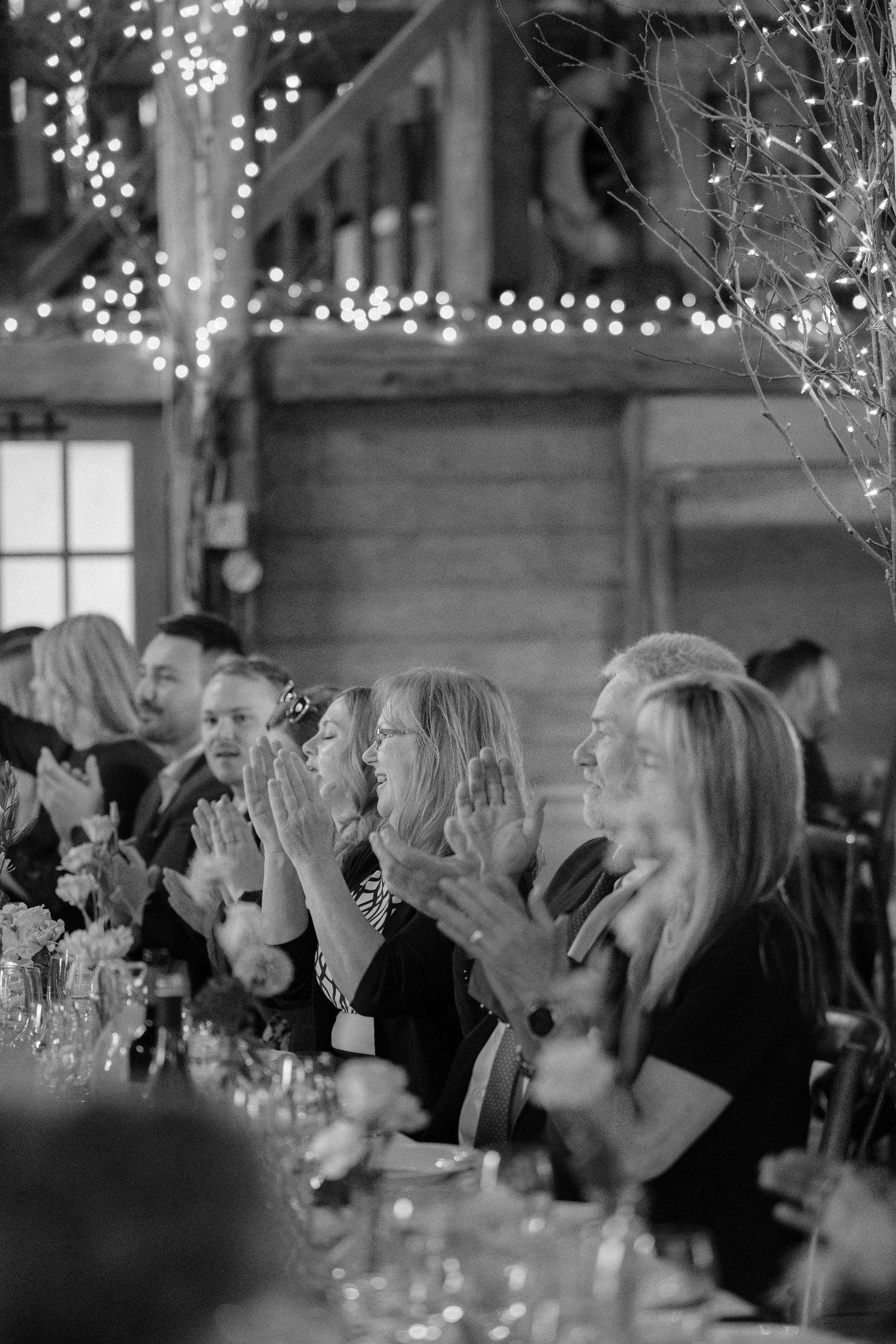 Wedding guests clap and laugh at long table adorned with fairy lights in rustic barn.