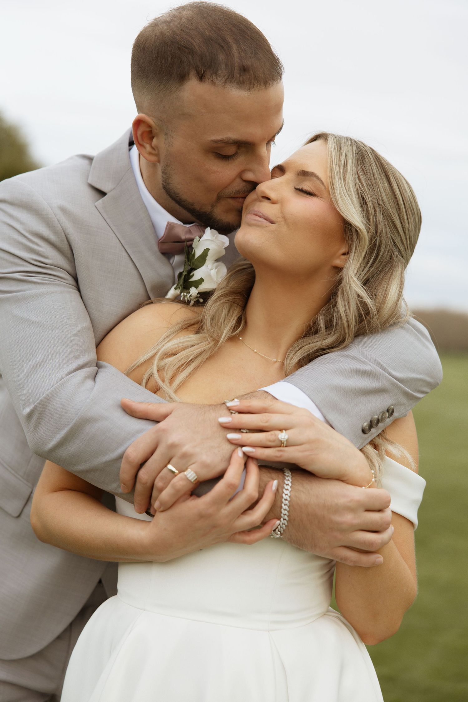 Bride and groom embrace outdoors, groom kissing bride's cheek, both wearing wedding rings, in gray suit and white gown.