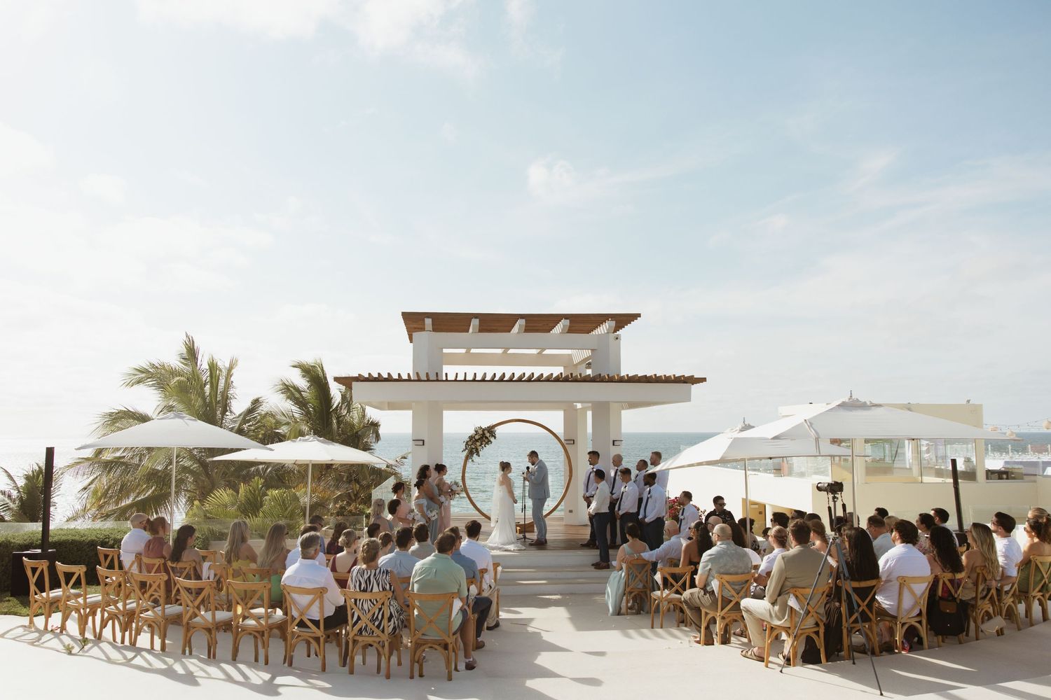 Outdoor beach wedding ceremony with guests seated on wooden chairs facing a white arch gazebo under a bright sky.