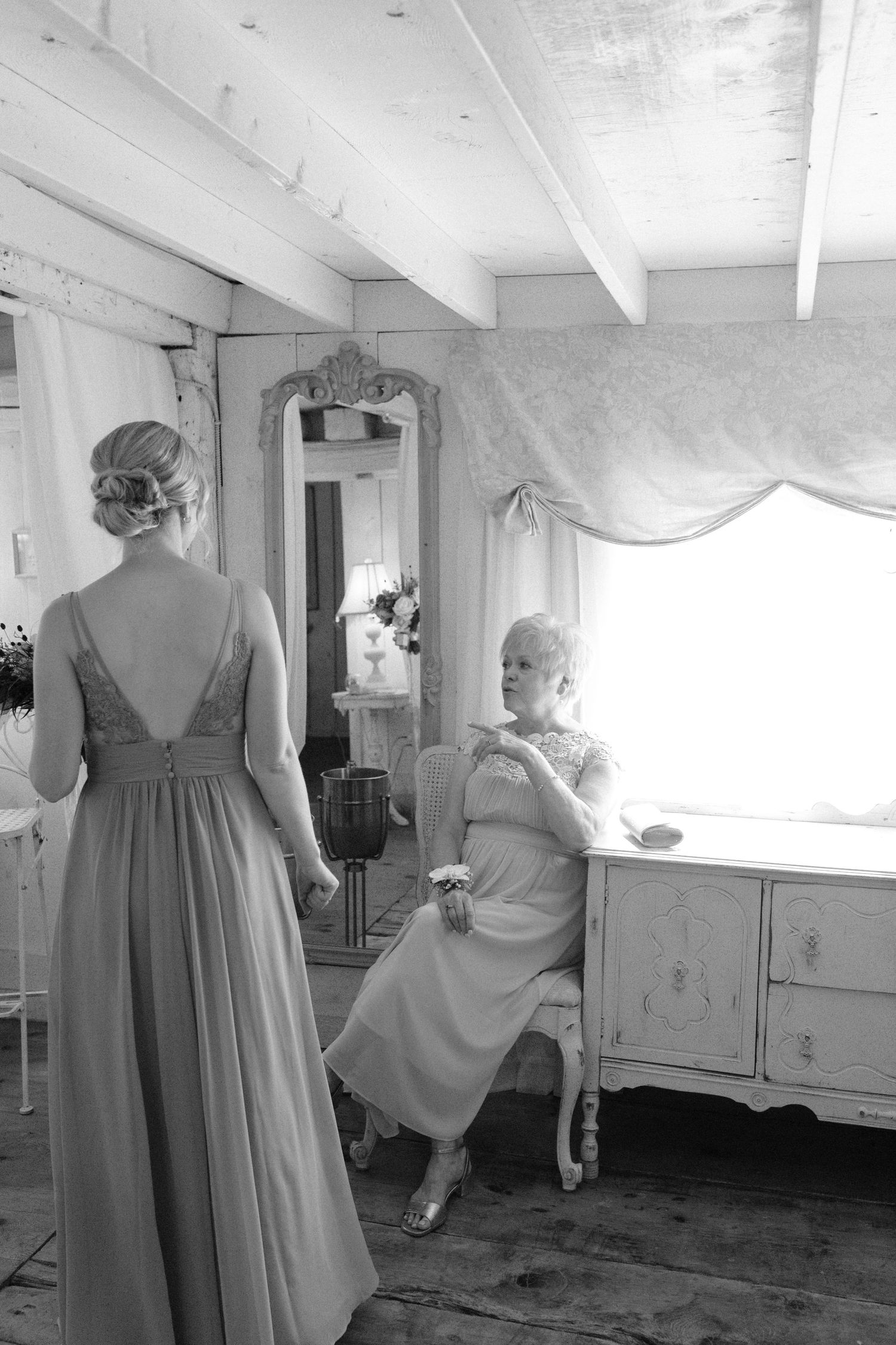 Bride in elegant gown viewed from behind as older woman sits by window in vintage dressing room.