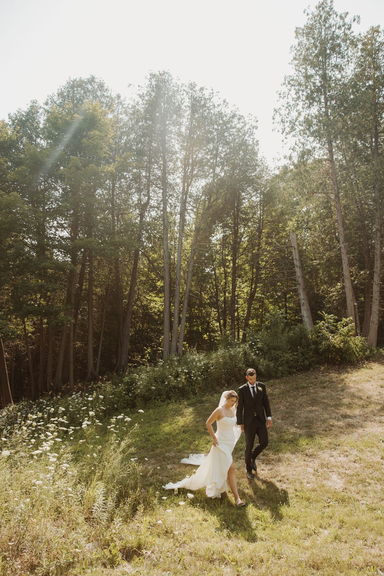 Bride and groom walking hand in hand through a sunlit forest clearing on their wedding day.