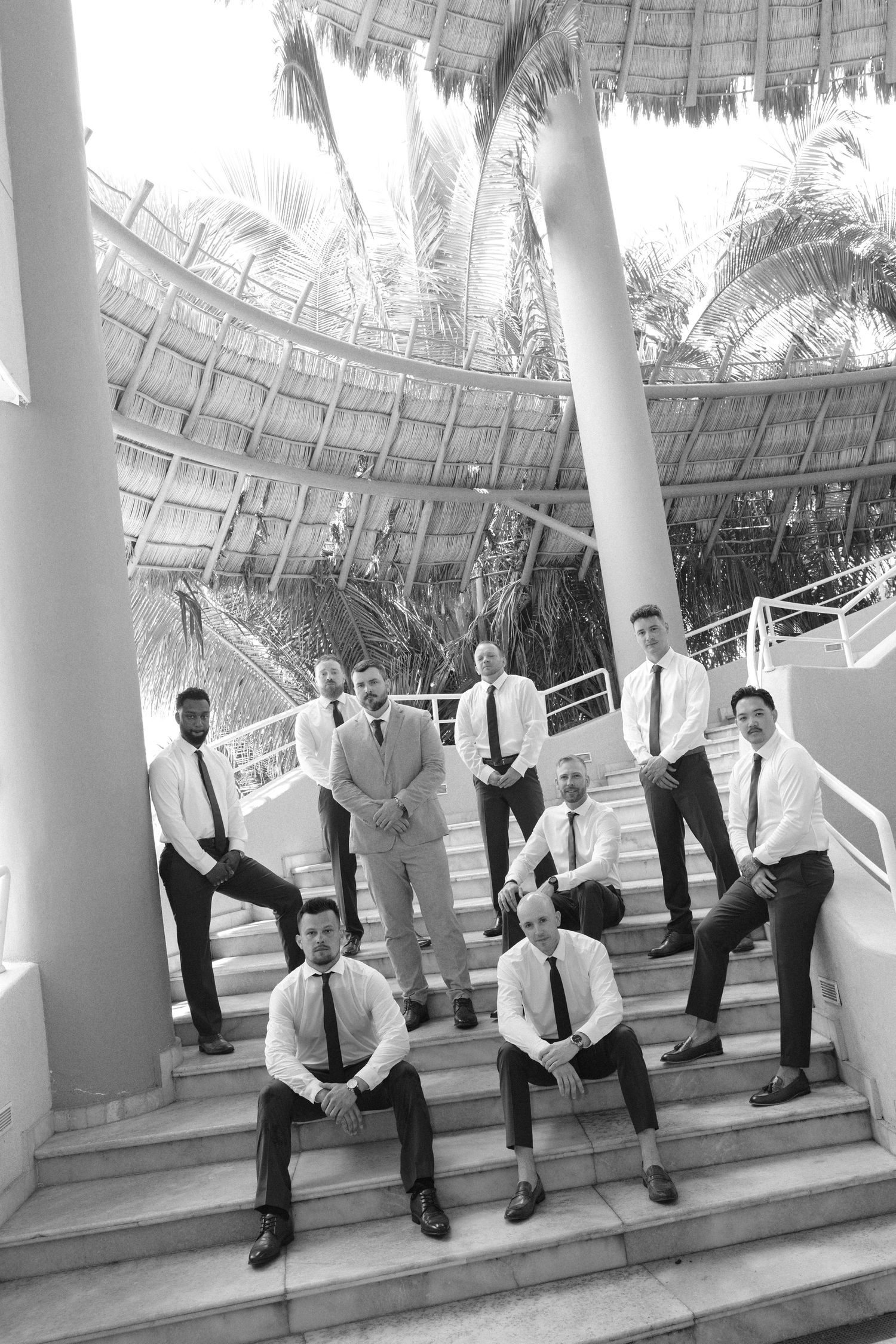 Black and white photo of groomsmen posing on outdoor stairs with palm trees and thatched roof at a tropical wedding venue.
