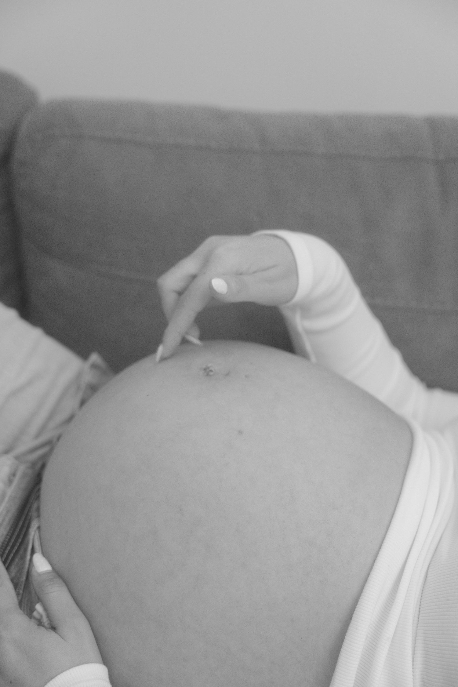 Black and white close-up of pregnant belly with hand gently resting on it against sofa.