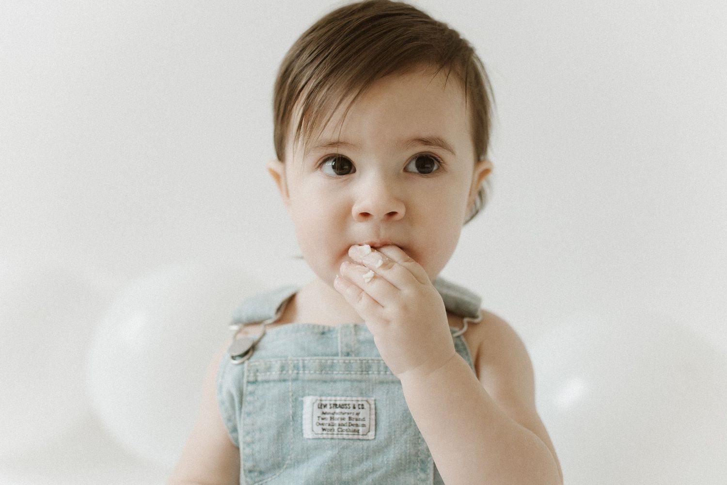 Adorable baby in denim overalls chewing on fingers while posing against a clean white background.