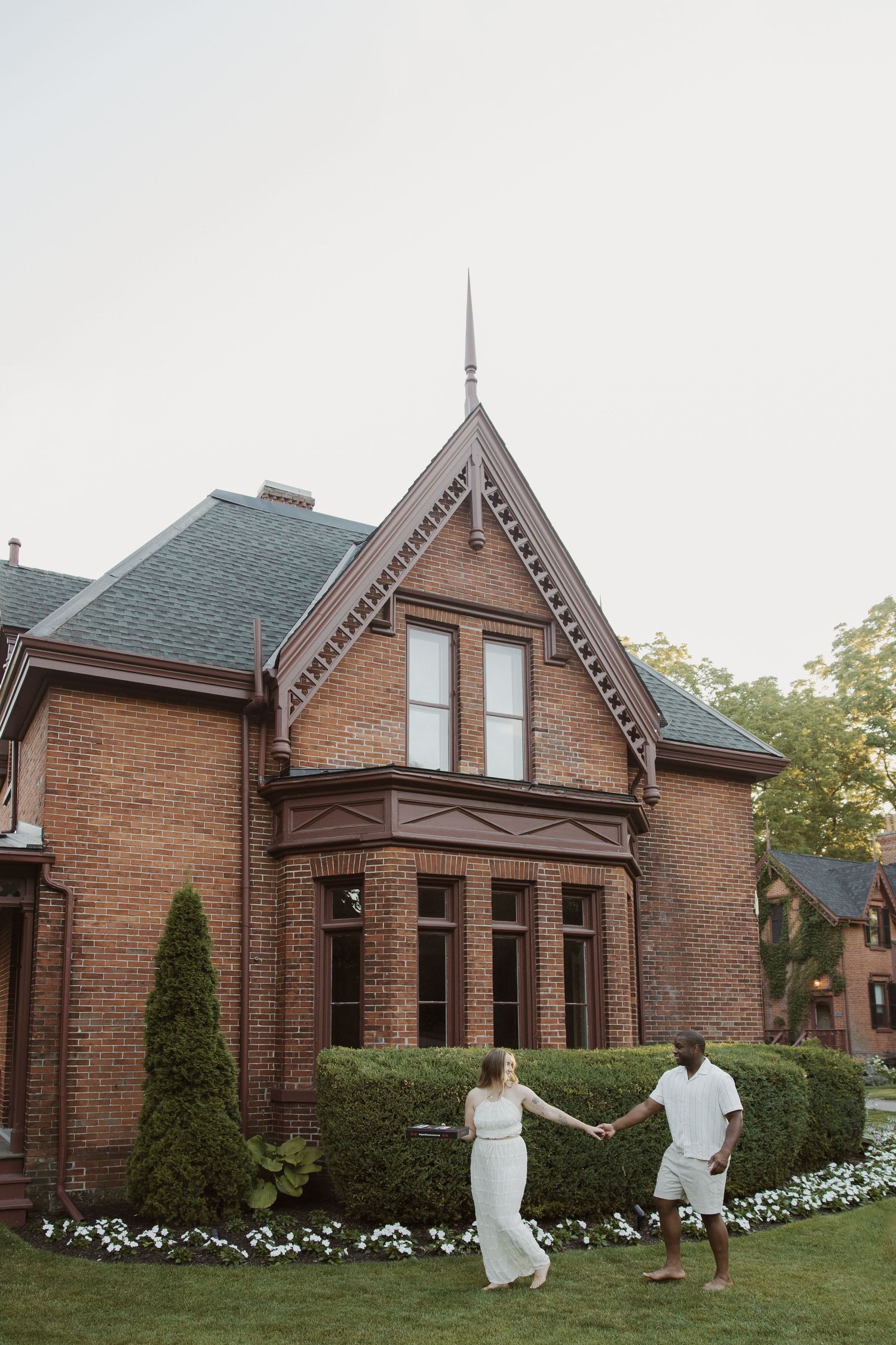 Couple holding hands on lawn in front of elegant brick Victorian mansion.